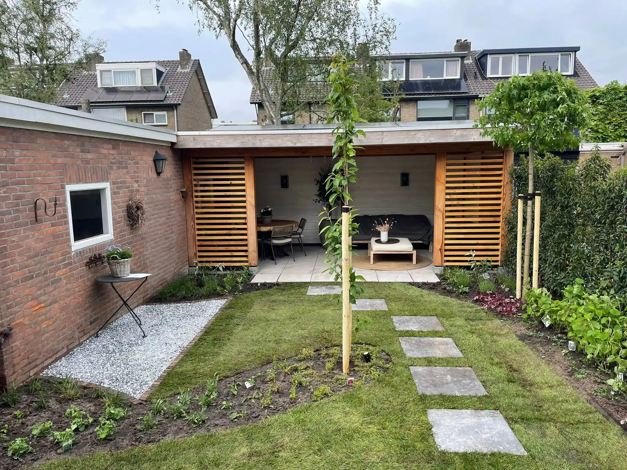 Achtertuin met een houten overkapping tussen twee lamellenwanden, een gazon met stapstenen, een vak wit grind langs de bakstenen muur en verse borders