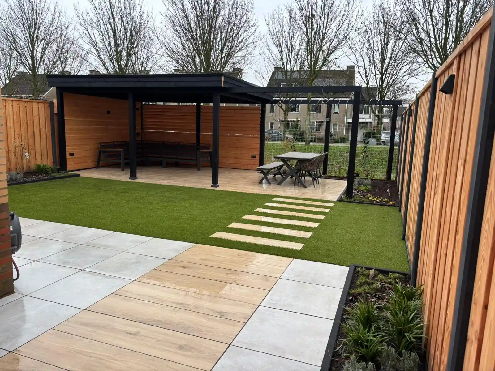 Achtertuin met een zwarte overkapping met houten wanden en ingebouwde banken, een veld kunstgras met stapstenen en een terras van houtlook tegels