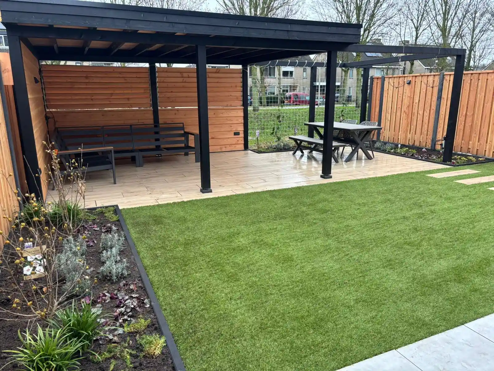 Zwarte overkapping met houten wanden en ingebouwde zitbanken, met daarnaast een pergola boven een eethoek en een ruim veld kunstgras