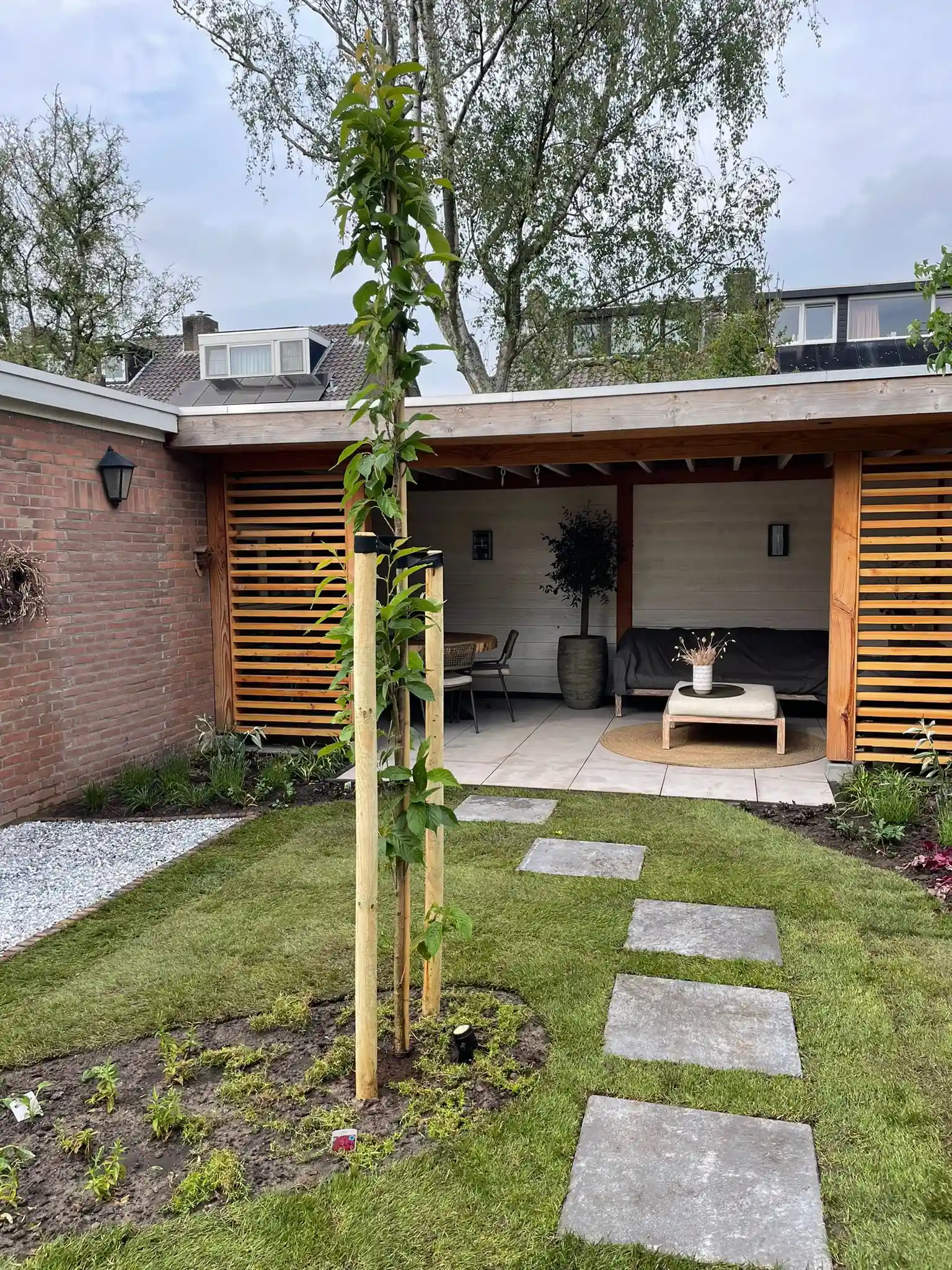 Houten overkapping met loungebank en eethoek achter openslaande lamellenwanden, met een gazon, een pad van grote stapstenen en een jonge boom met boompalen ervoor