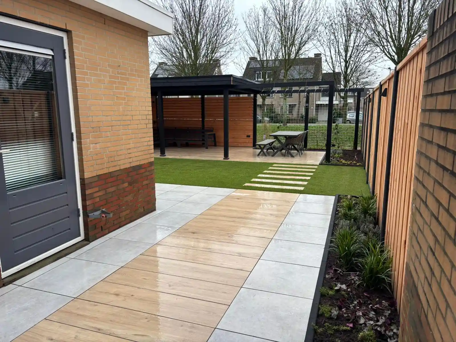 Terras van lichte houtlook tegels langs de gevel van een bakstenen woning, met een border met heuchera langs de schutting en de overkapping op de achtergrond