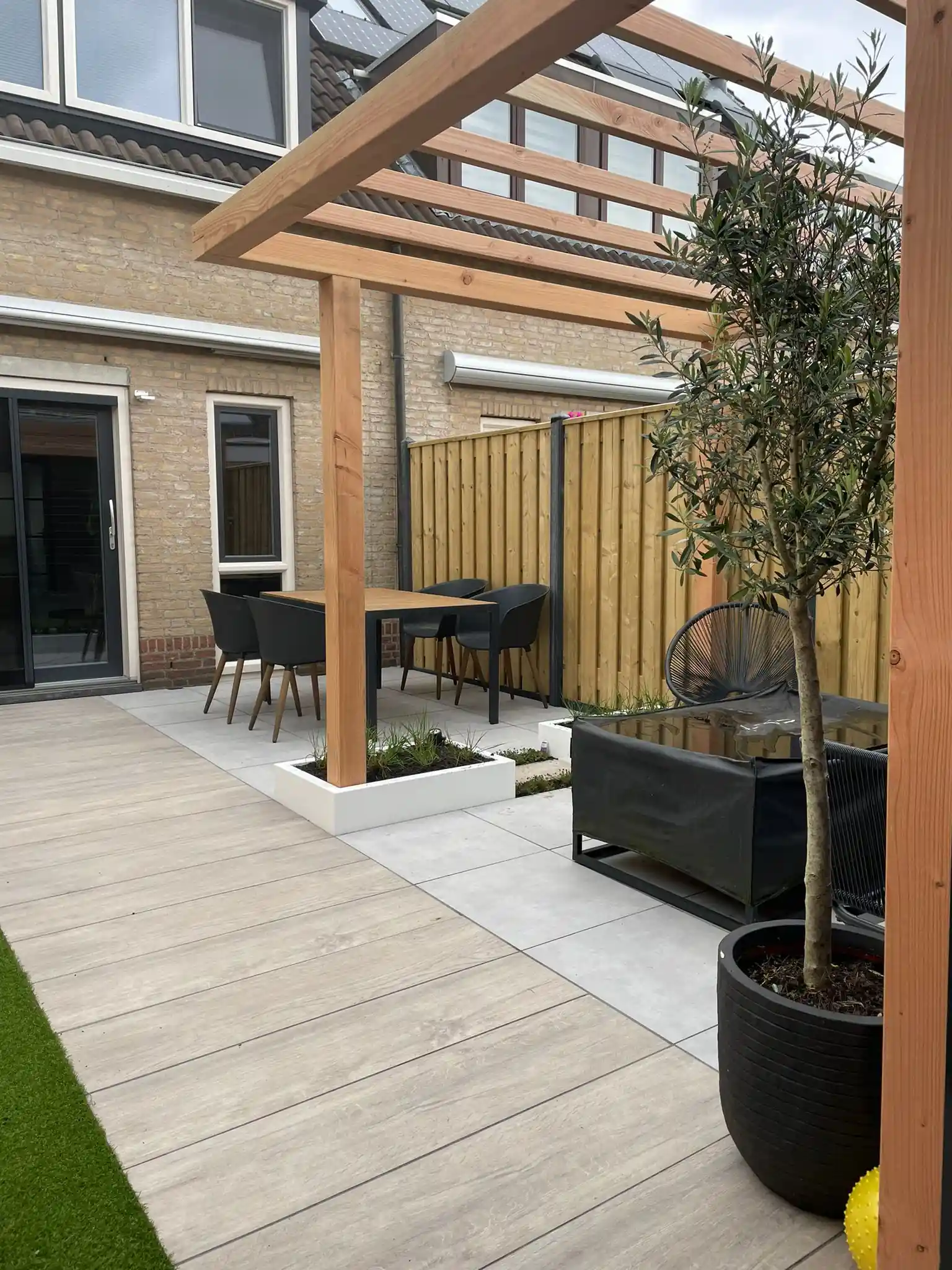 Eettafel met zwarte kuipstoelen onder een Douglas pergola langs de gevel, met een witte plantenbak met siergrassen en een olijfboom in pot op de voorgrond