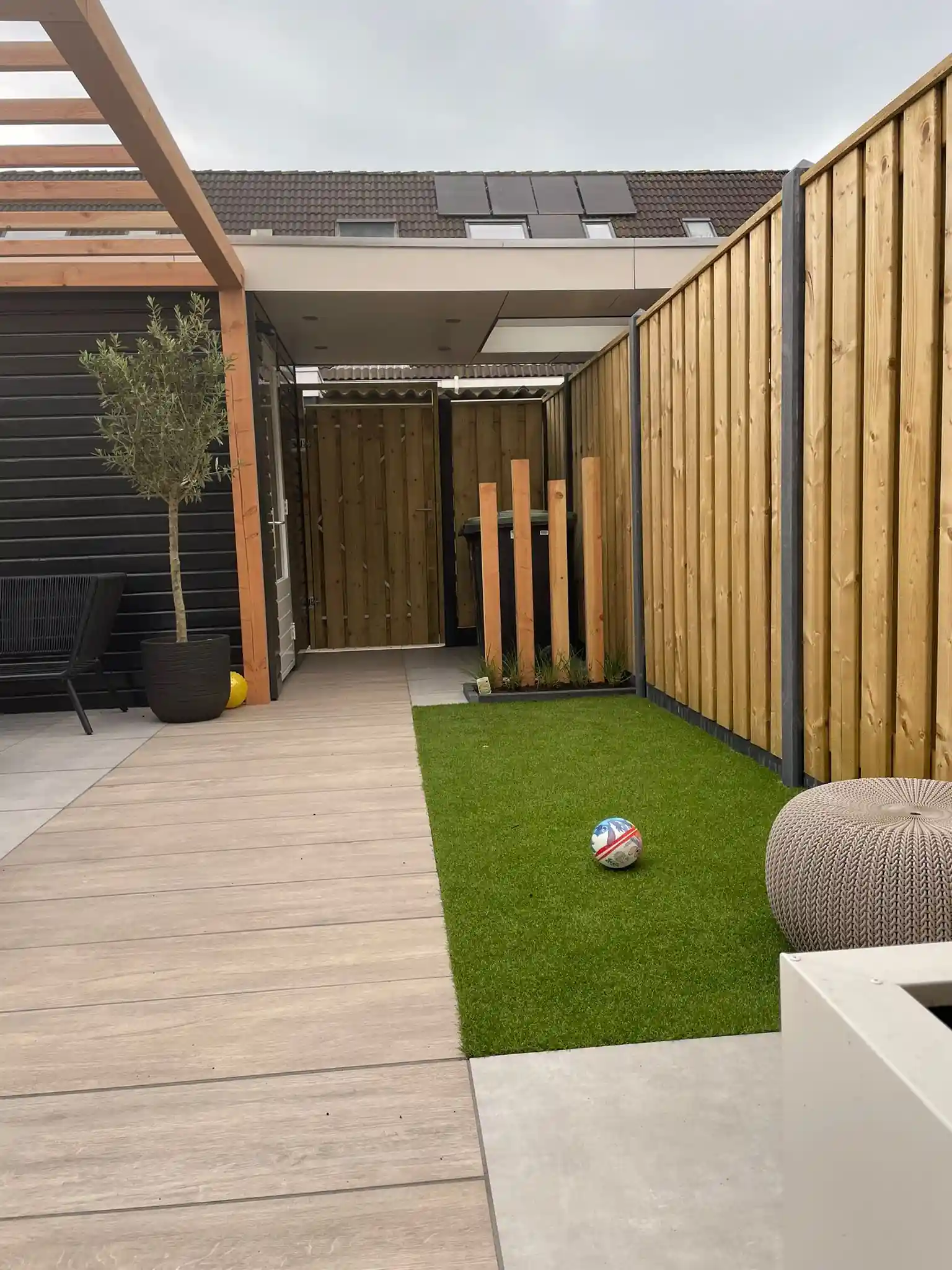 Strook kunstgras naast een terras van houtlook keramiek, met een houten schutting, staande Douglas palen en een olijfboom in een donkere pot