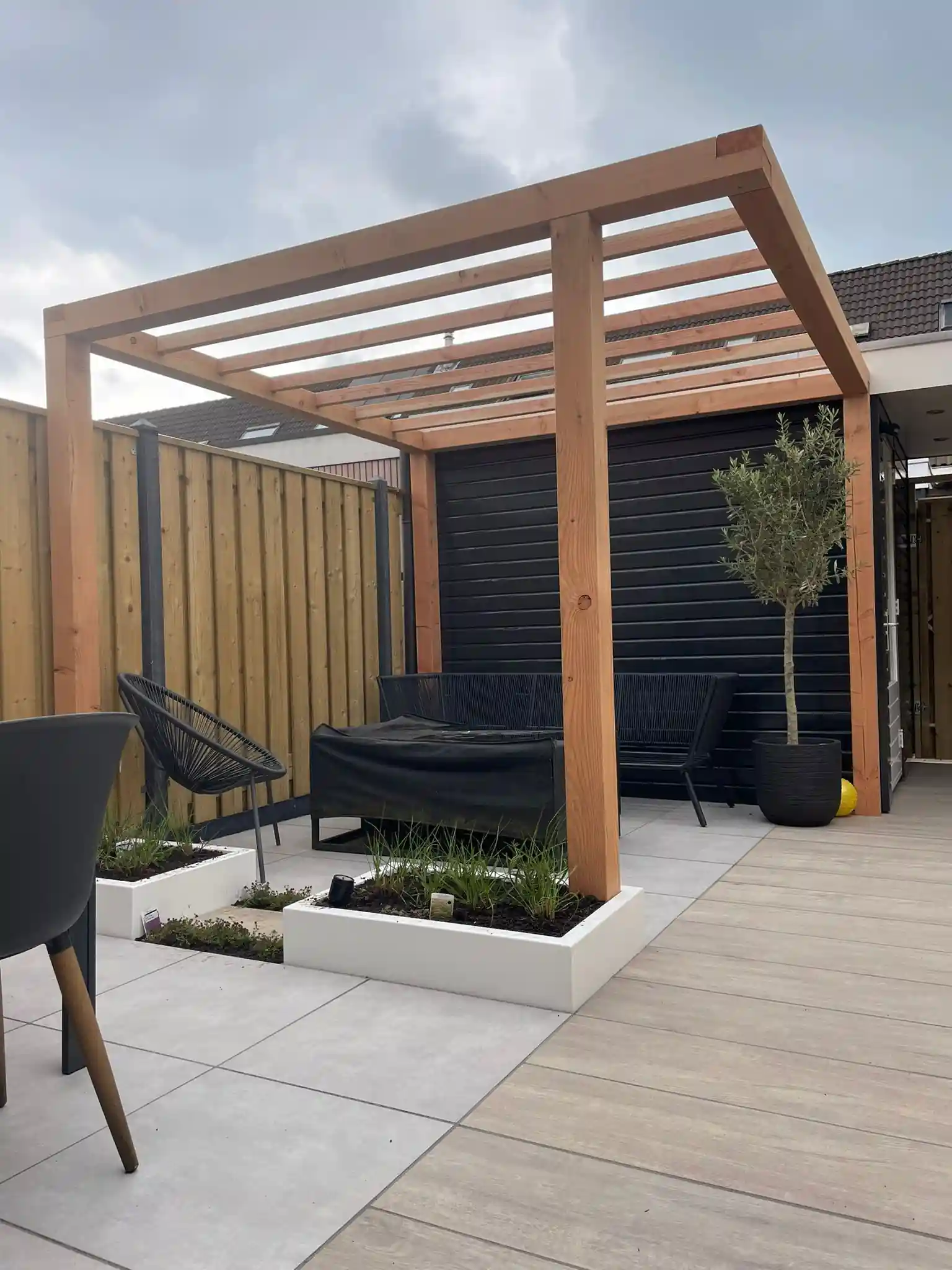 Douglas pergola boven een loungehoek met zwarte tuinmeubelen tegen een donkere houten wand, met een olijfboom in pot en een witte plantenbak met siergrassen