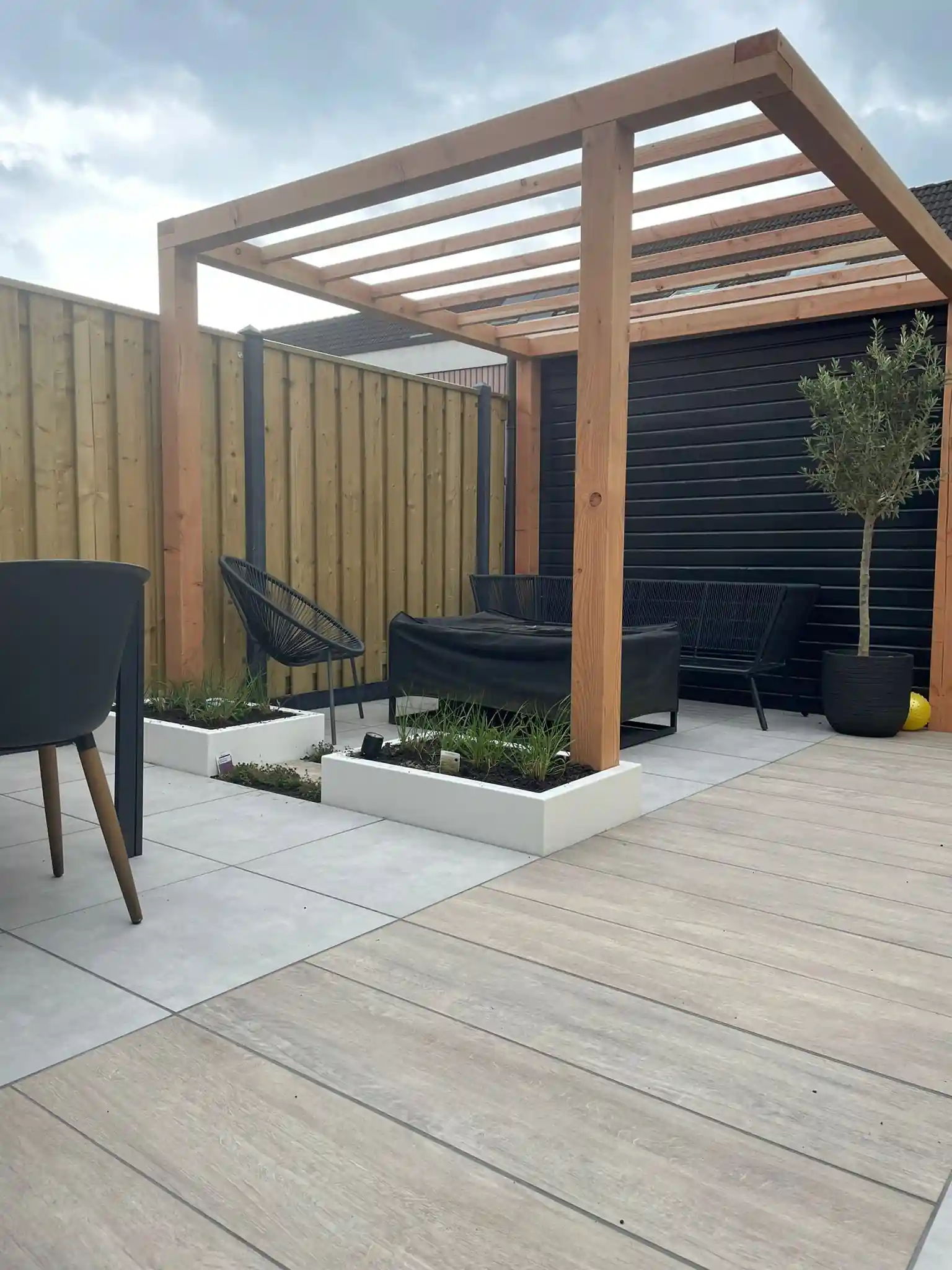 Terras van grijze tegels en houtlook keramiek onder een Douglas pergola, met lage witte plantenbakken met siergrassen en zwarte tuinmeubelen