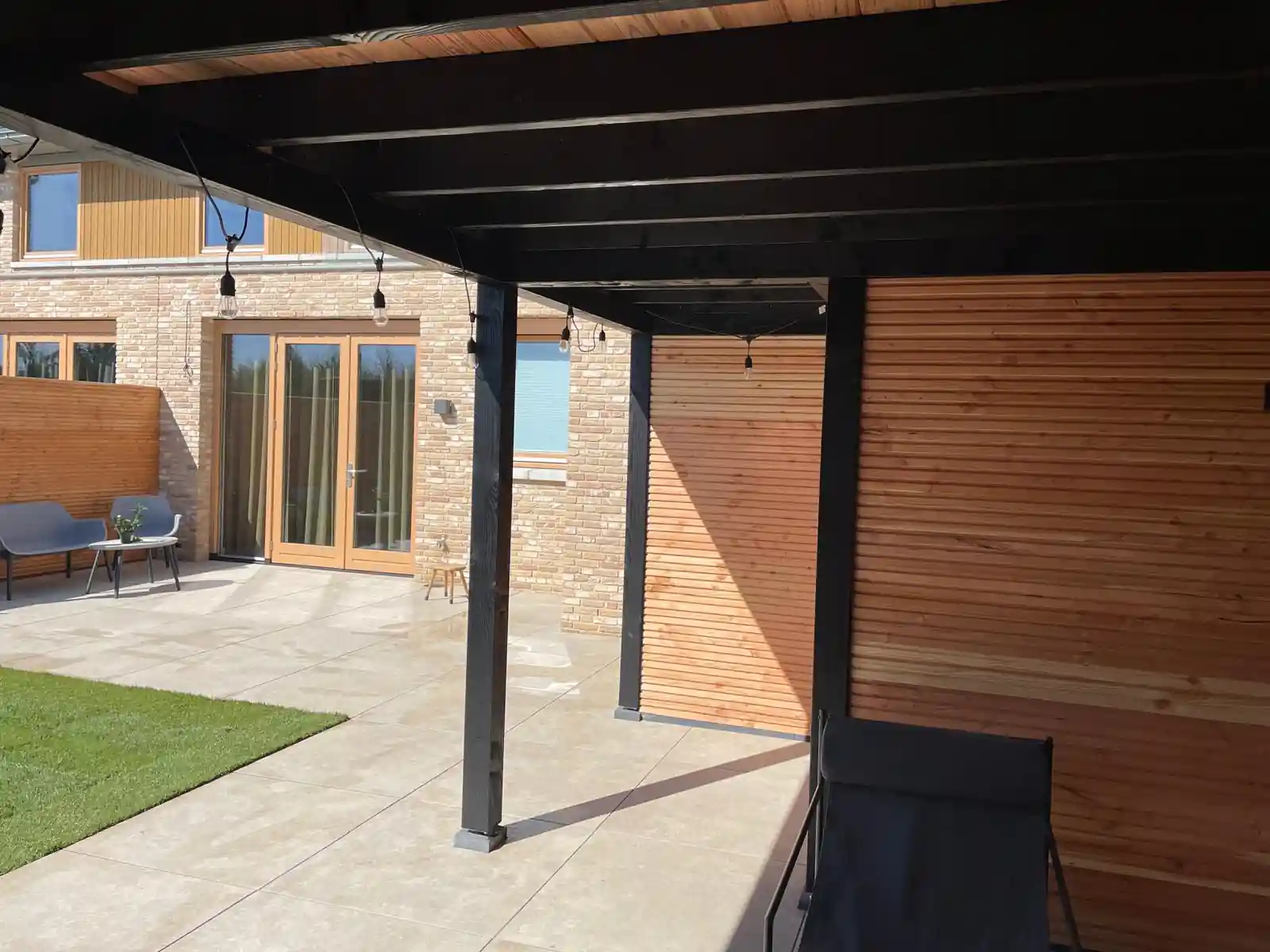Terras van lichte tegels onder de overkapping met een lichtsnoer tussen de balken, met zicht op de woning en een strook kunstgras