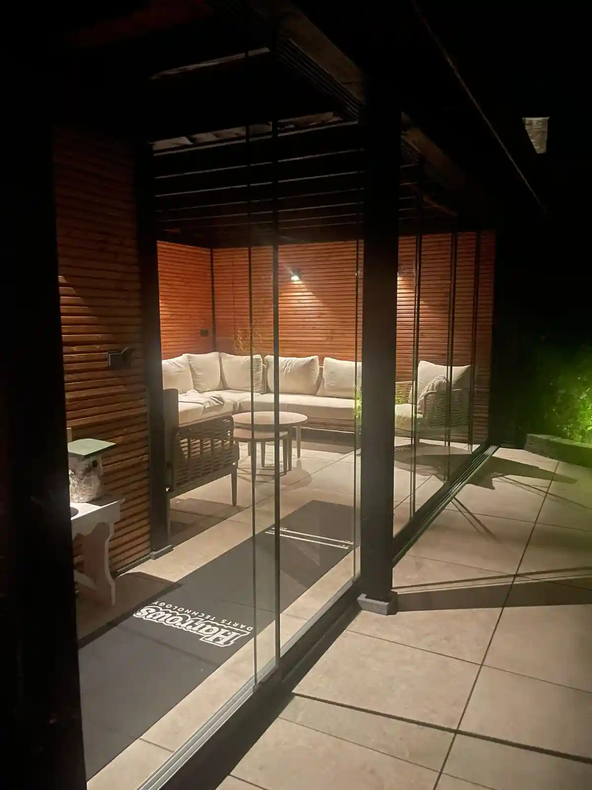 Verlichte loungehoek met lichte hoekbank en ronde bijzettafels in de tuinkamer, gezien door de glazen schuifwand vanaf het terras