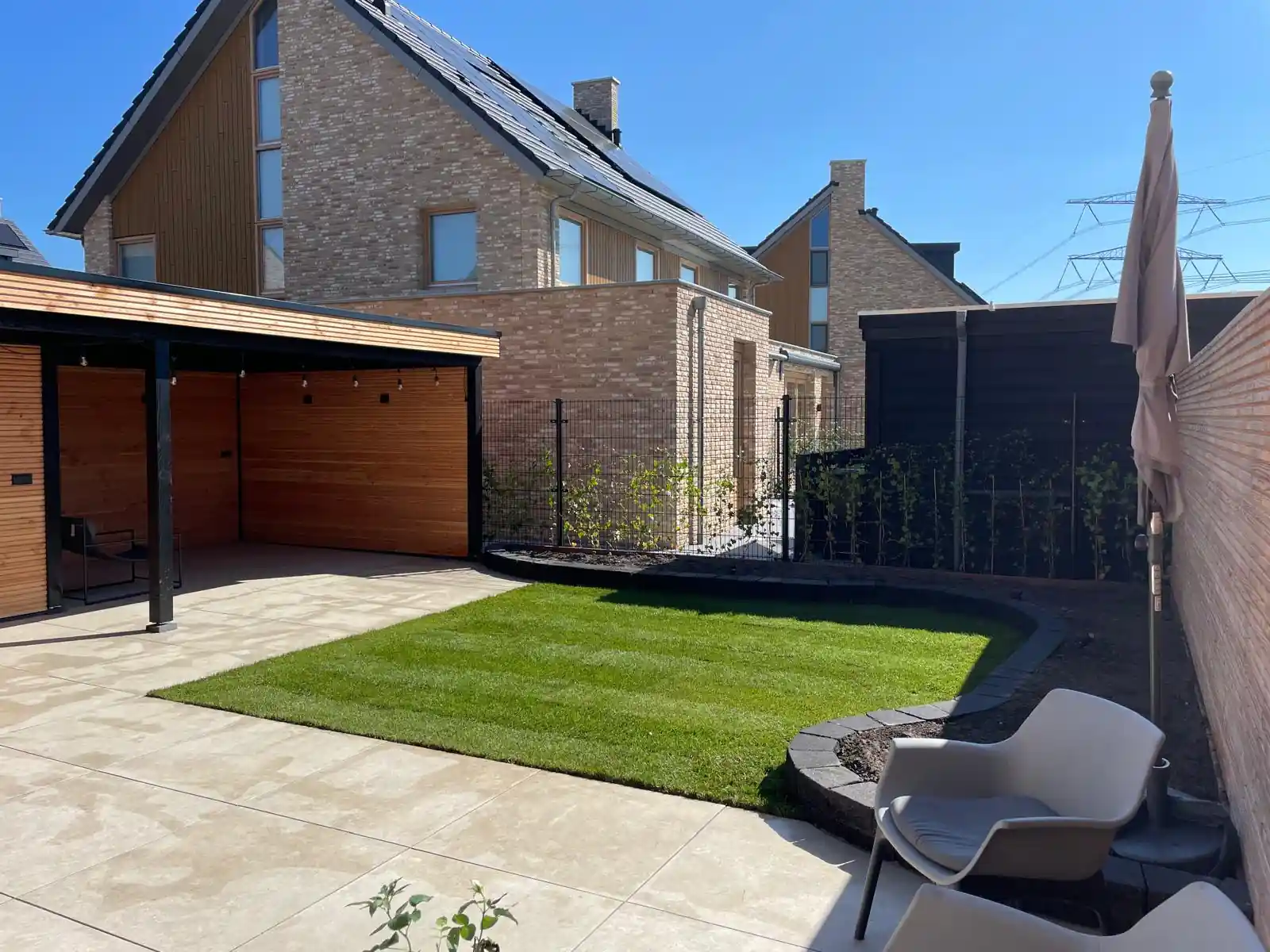 Achtertuin met een donkere overkapping met houten wanden, een rechthoekig gazon met een gebogen border van stapelblokken en een ruim terras van lichte tegels