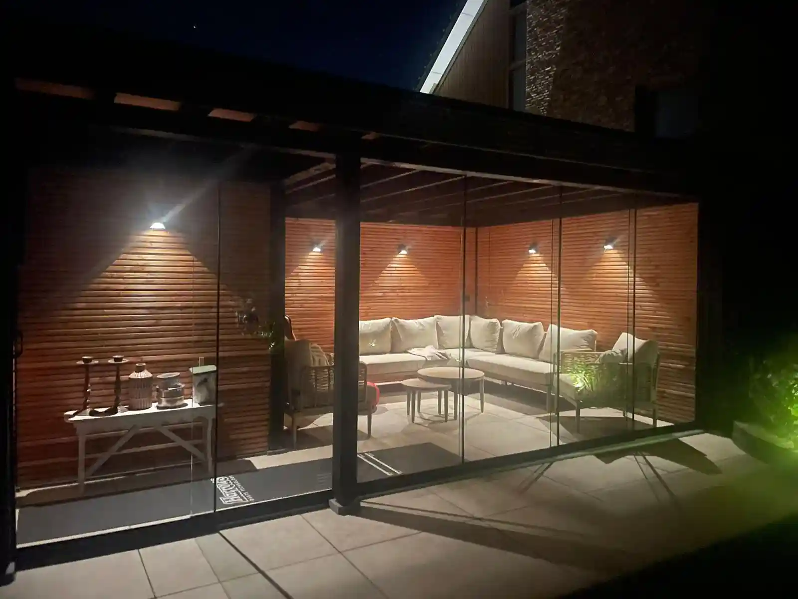 Tuinkamer in de avond met wandspots die lichtkegels op de Douglas wanden werpen, met een lichte hoekbank achter de glazen schuifwanden