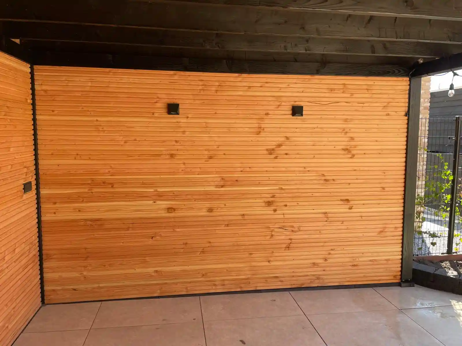 Binnenwand van de overkapping met Douglas rabatdelen en twee zwarte wandspots, onder een dakconstructie met donkere balken