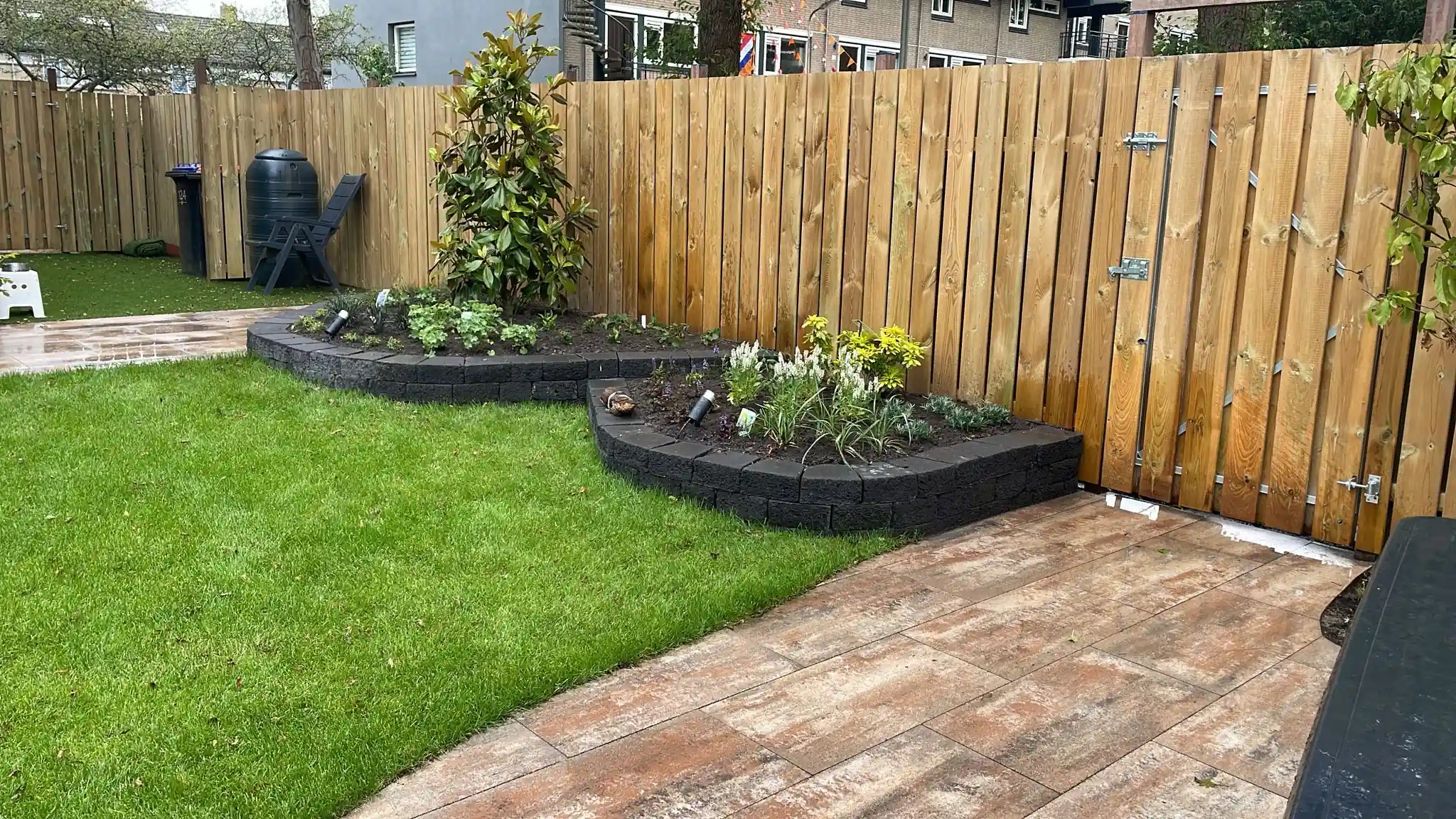 Verhoogde border van lavablokken met witte salie en grondspots naast een houten poort in de schutting, met een gazon en een pad van houtlook tegels