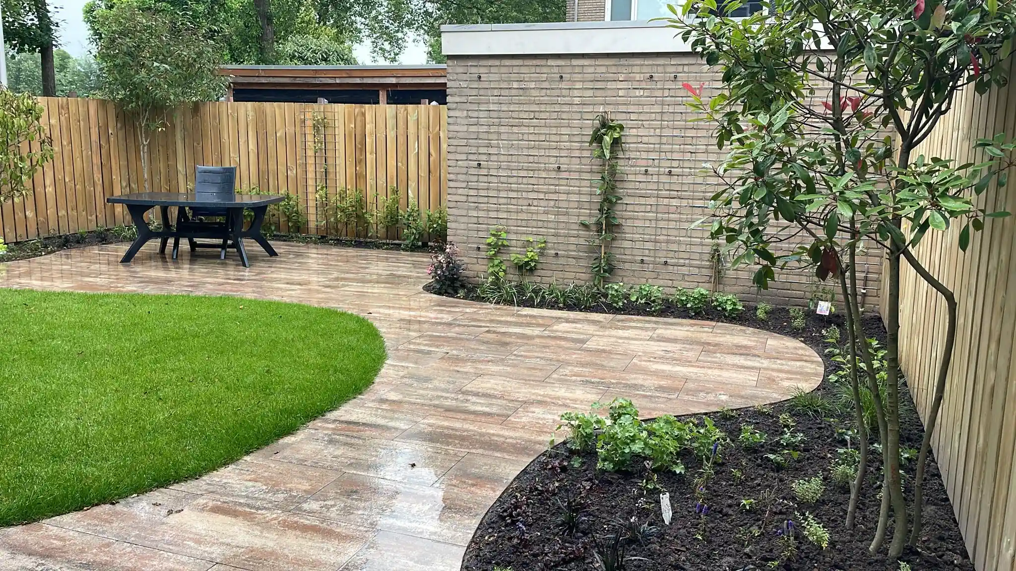 Gebogen terras van houtlook tegels rond een ovaal gazon, met een eethoek bij de schutting en jonge klimplanten tegen een lichte bakstenen muur