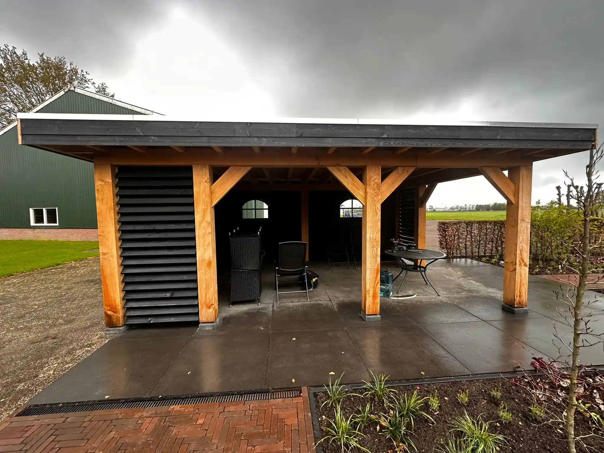 Vrijstaande eikenhouten overkapping met zwart plat dak en lamellenwanden, met een tegelvloer, tuinset en een beukenhaag met zicht op het open veld