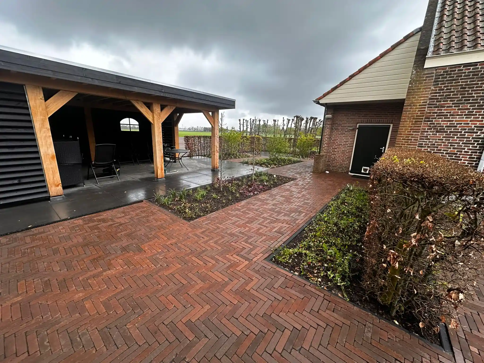 Eikenhouten overkapping met zwarte lamellenwand naast een ruim terras van roodbruine waalformaat klinkers in visgraatverband, met verse borders en uitzicht op het weiland