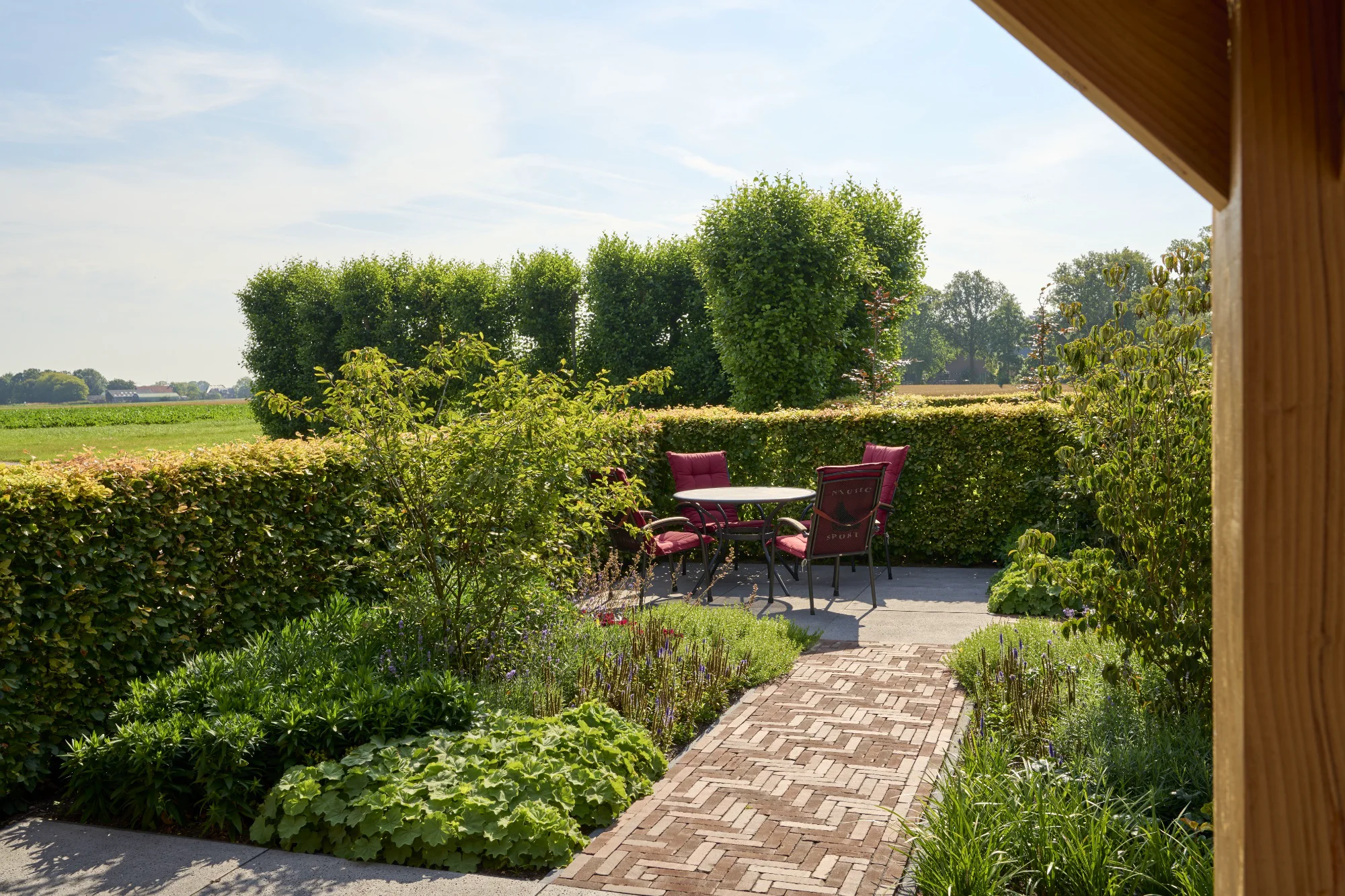 Zitje met ronde tafel en rode kussens op een tegelterras, bereikbaar via een klinkerpad tussen weelderige borders met vaste planten en beukenhagen