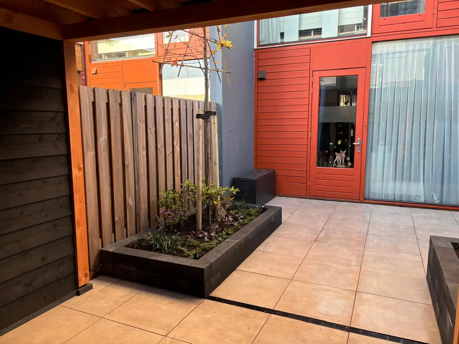 Overzicht van de compacte achtertuin met een terras van lichte tegels, een verhoogde border met amberboom en beplanting, en de rode gevel van de woning