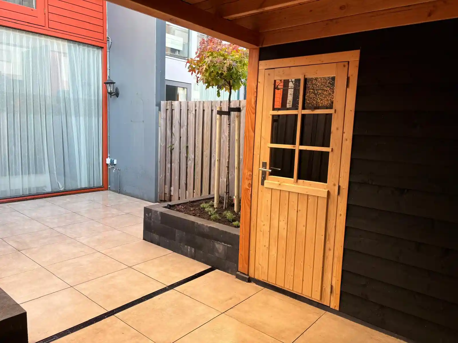 Terras van lichte tegels met een lijngoot voor de pui van de woning, met een verhoogde border met amberboom en de Douglas schuurdeur rechts