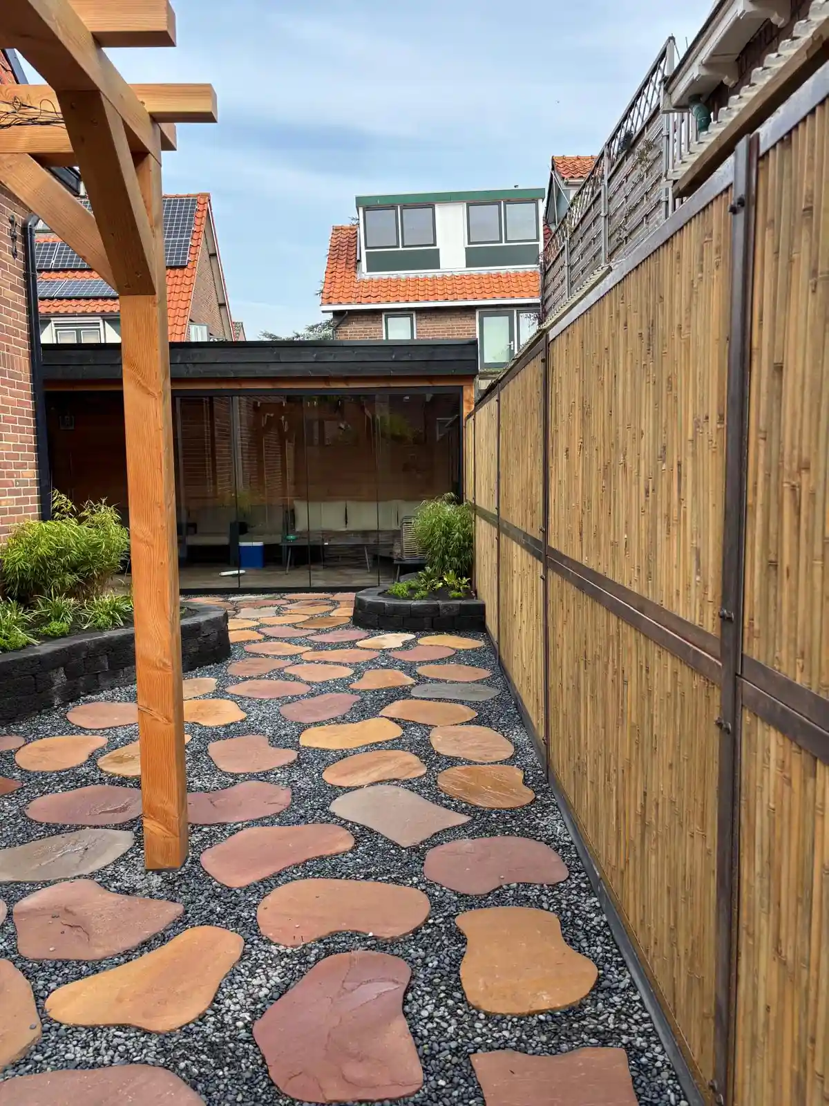 Hoge bamboeschutting met roestbruine staanders langs een terras van flagstones in split, met een pergola en de tuinkamer op de achtergrond