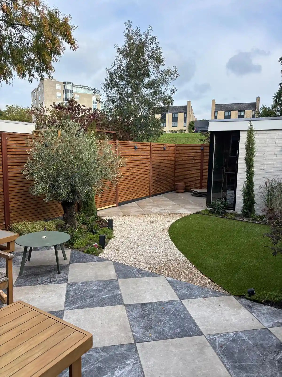 Terras in dambordpatroon van grijze en lichte tegels met een olijfboom in de hoek, een grindpad naar een rond veld kunstgras en een witte tuinkamer