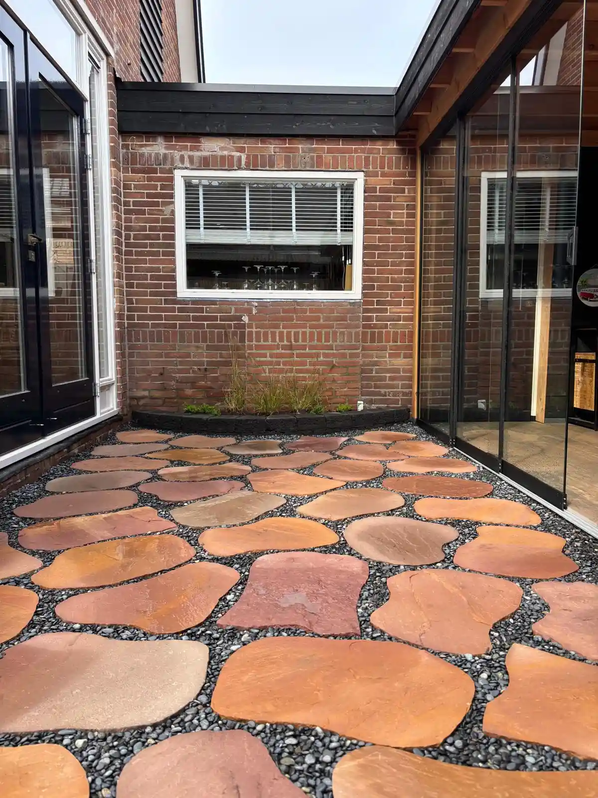 Terras van roodbruine flagstones in grijs split langs een rode bakstenen gevel met wit raam, met siergrassen in een border van zwarte stapelblokken