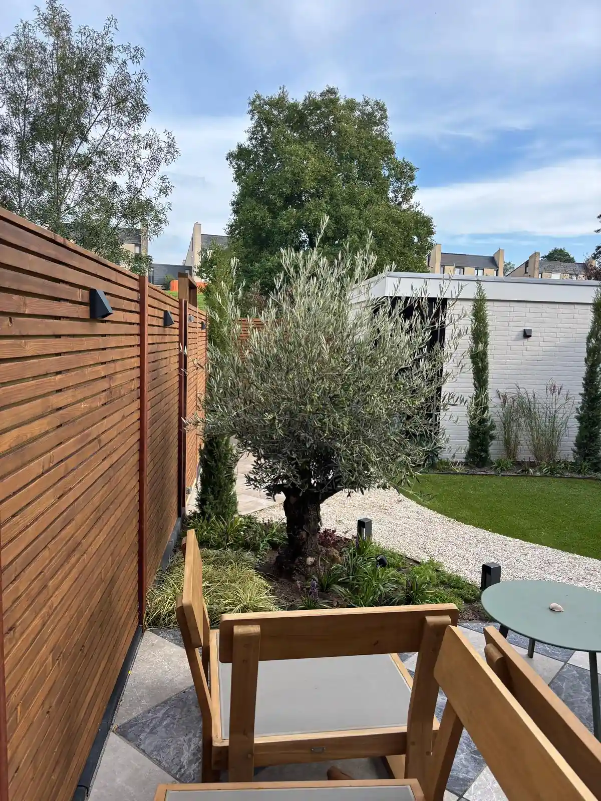 Hardhouten lattenschutting met wandlampen langs een zithoek met houten stoelen, met zicht op de olijfboom, het grindpad en het kunstgras