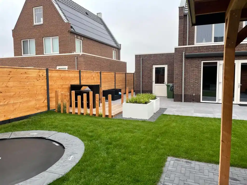 Ruim gazon met een verdiepte trampoline, met een vlonder met statafel achter Douglas paaltjes en een witte plantenbak met siergrassen langs het terras