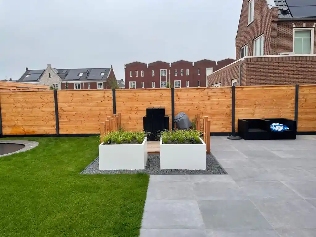 Twee witte plantenbakken met siergrassen en een houten zitrand ertussen, op een strook split langs een terras van grijze tegels en een Douglas schutting
