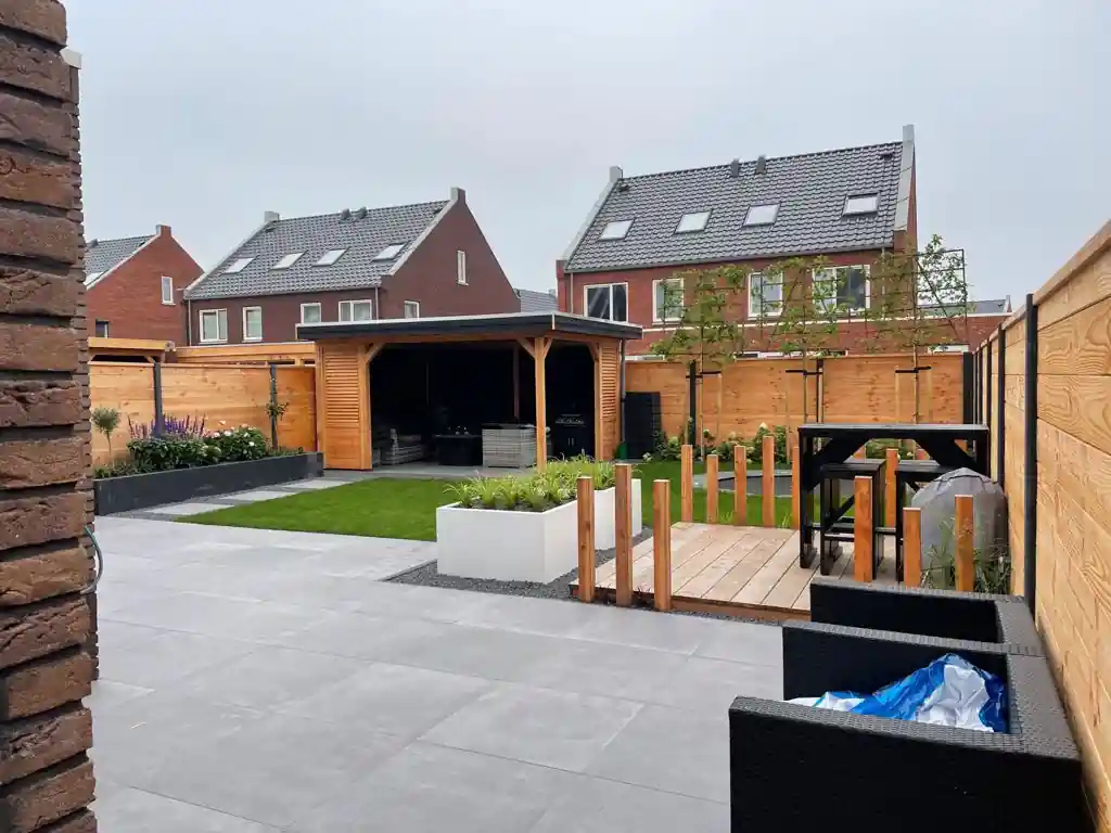 Ruim terras van grote grijze tegels met zicht op de overkapping, het gazon, de vlonder met statafel en jonge leibomen langs de Douglas schutting