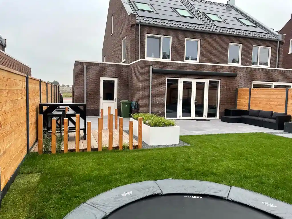 Houten vlonder met een zwarte statafel en krukken achter een rij Douglas paaltjes, met een witte plantenbak, gazon en de openslaande deuren van de woning