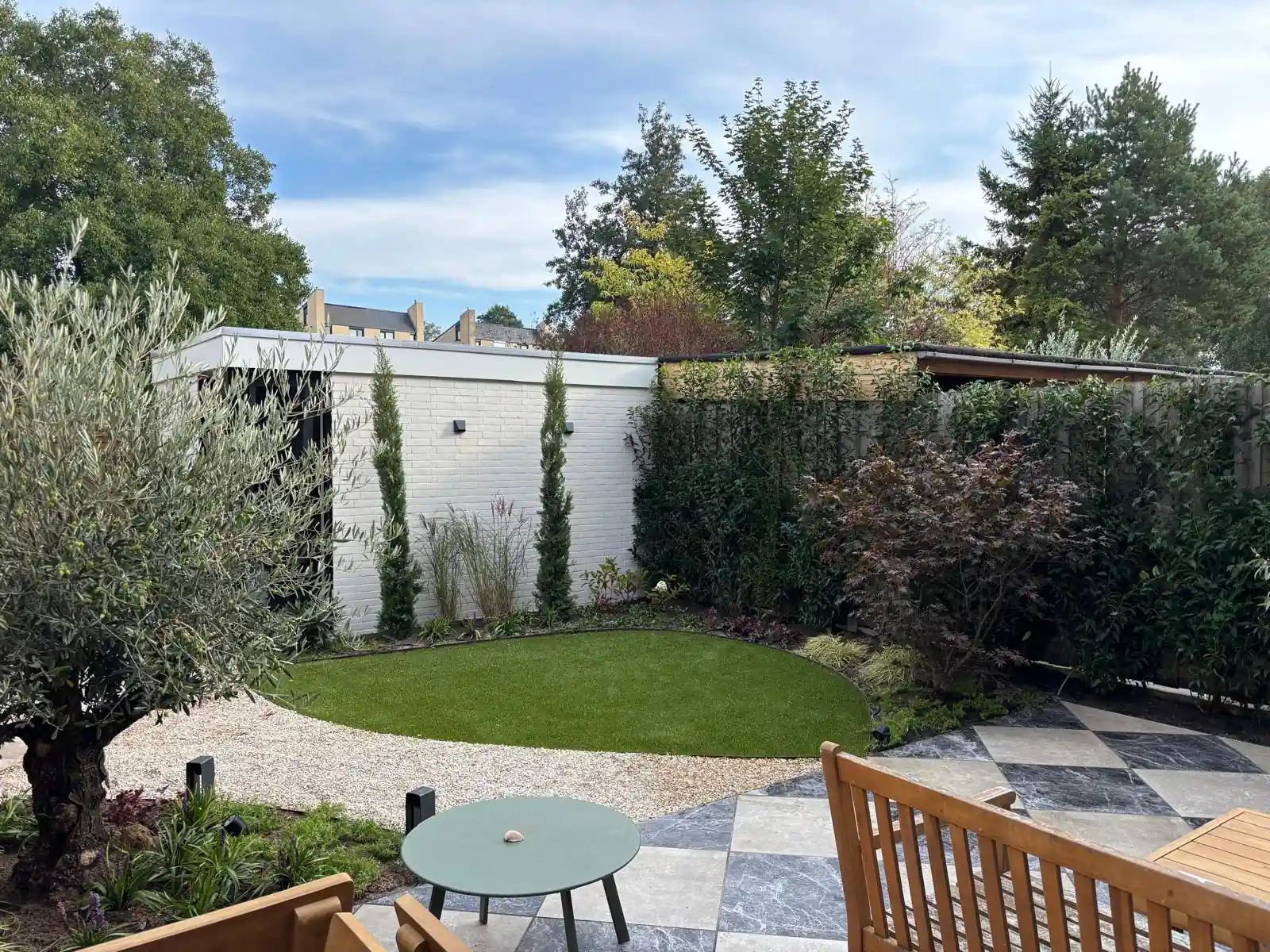 Overzicht van de achtertuin met een rond veld kunstgras, grindpad, witte tuinkamer met cipressen en een terras in dambordpatroon met houten meubels