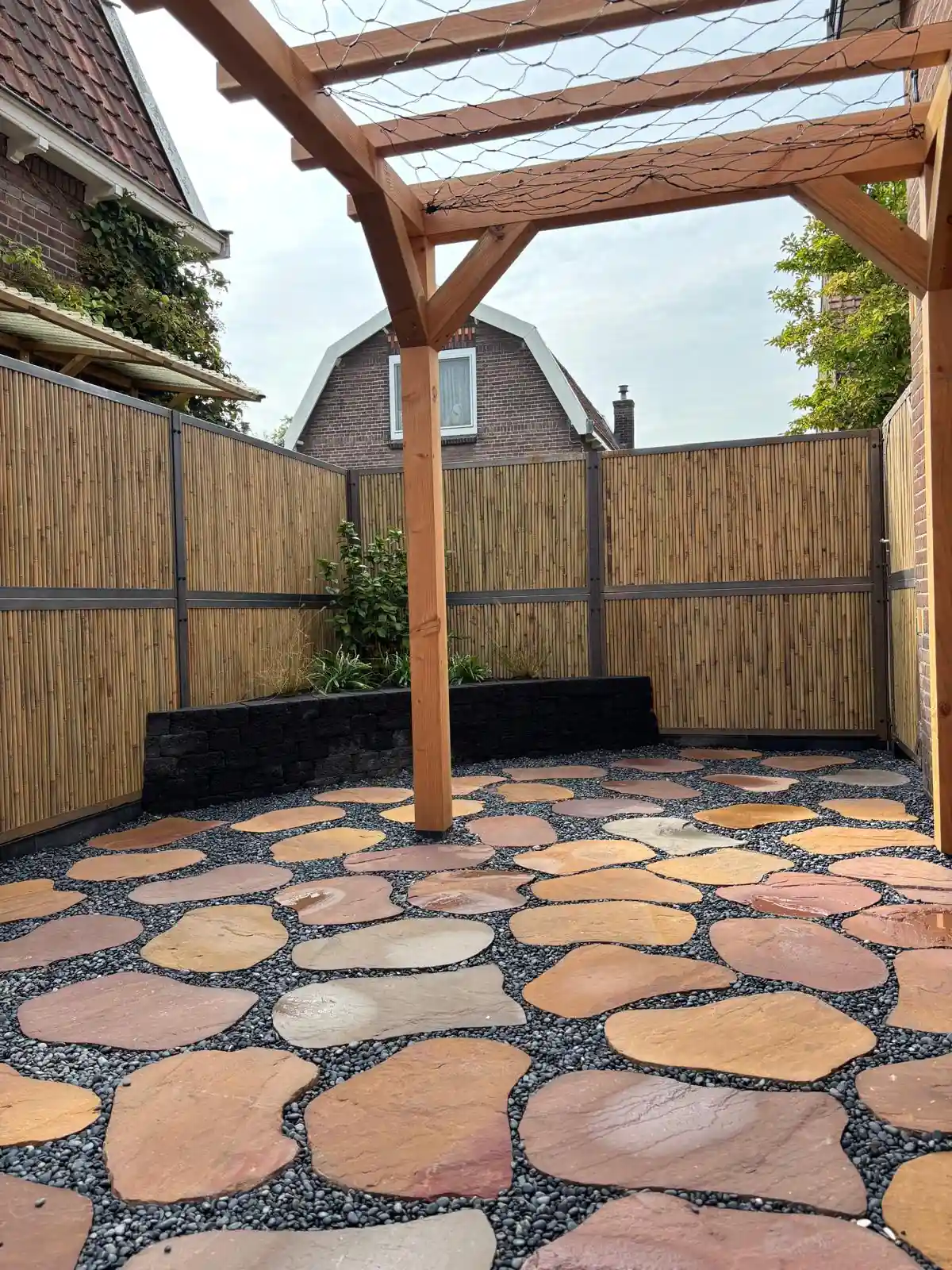 Douglas pergola boven een terras van roodbruine flagstones in grijs split, met een bamboeschutting en een verhoogde border van zwarte stapelblokken