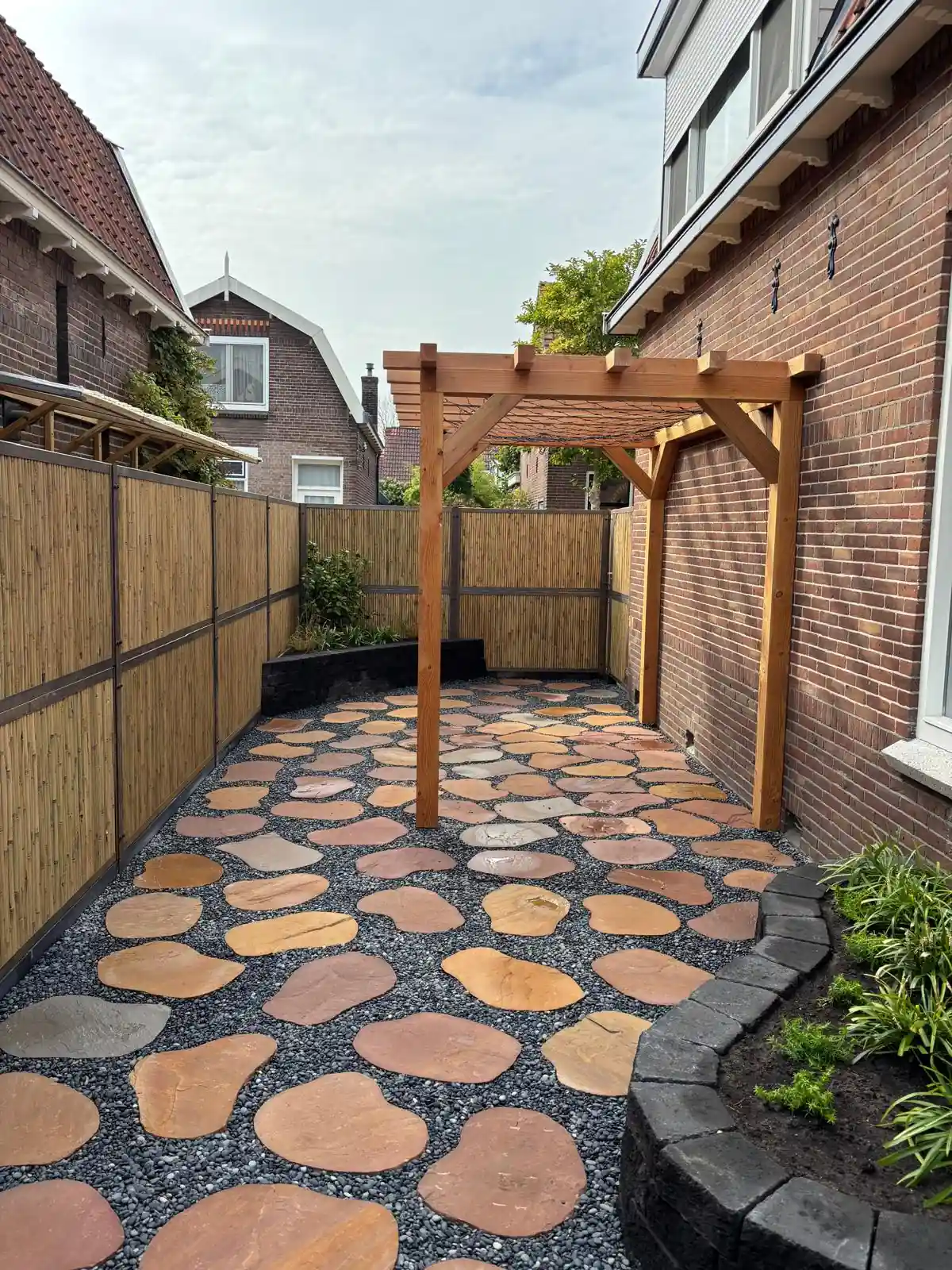 Douglas pergola tegen een bakstenen gevel, met een border van zwarte stapelblokken en een terras van flagstones in grijs split