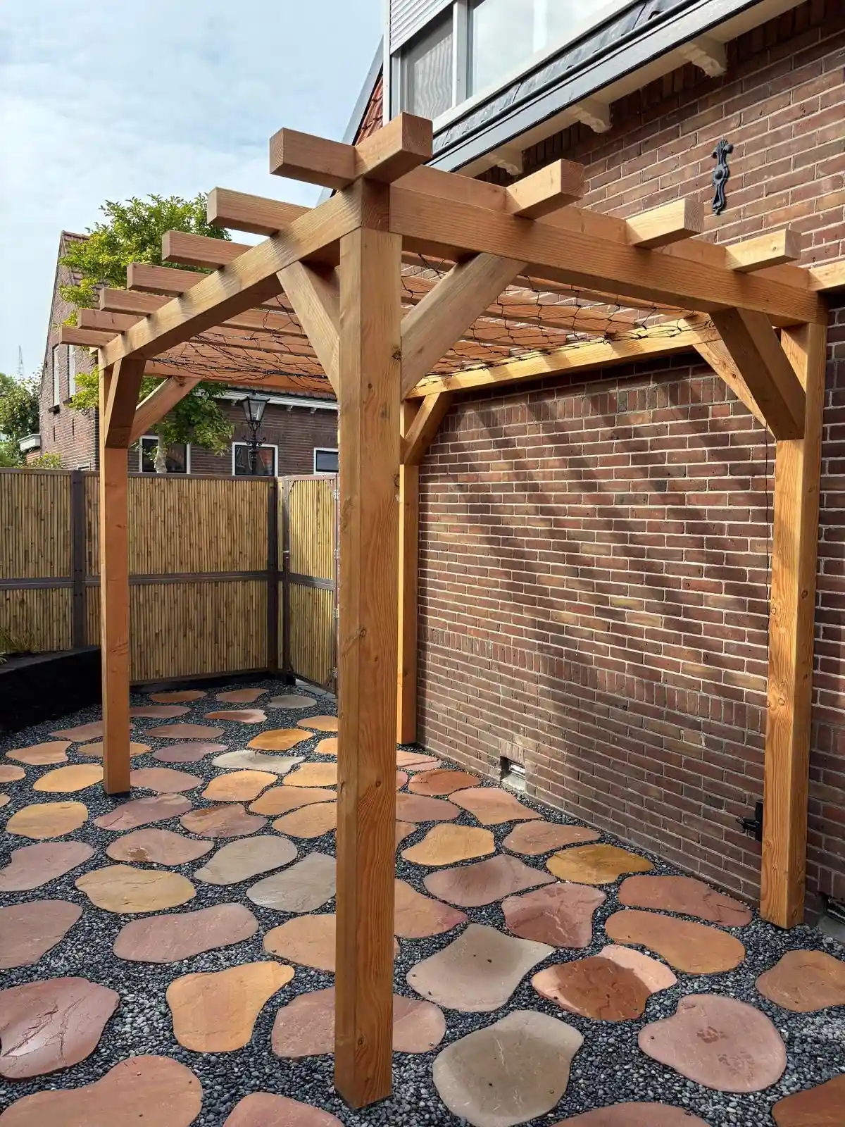 Vrijstaande Douglas pergola met gekruiste kopse balken tegen een rode bakstenen muur, met een terras van flagstones in split eronder
