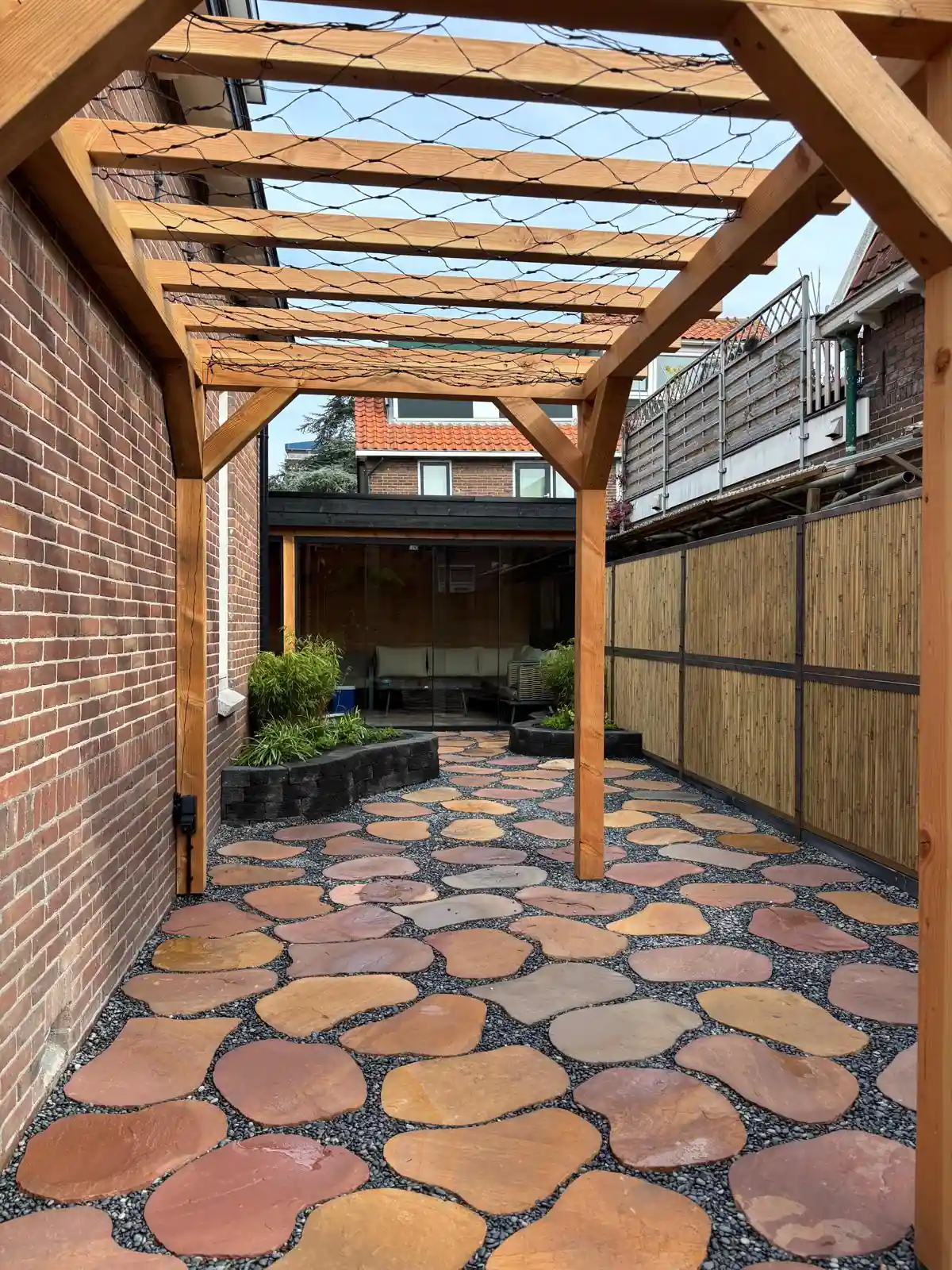 Terras van roodbruine flagstones in grijs split onder een Douglas pergola met klimnet, met bamboeschutting rechts en de tuinkamer op de achtergrond