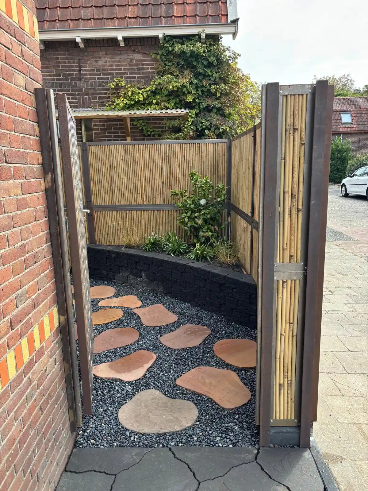 Open stalen poort met bamboevulling die uitkijkt op een pad van losse flagstones in grijs split, langs een verhoogde border van zwarte stapelblokken