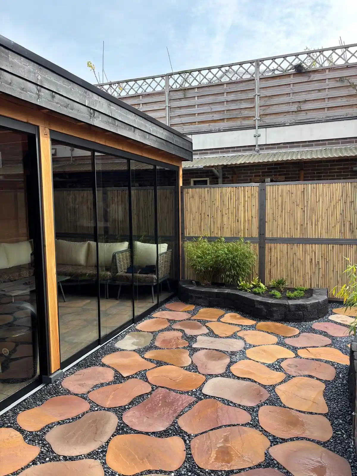 Tuinkamer met zwarte glazen schuifwanden en een rieten loungeset, met daarvoor een terras van roodbruine flagstones in grijs split