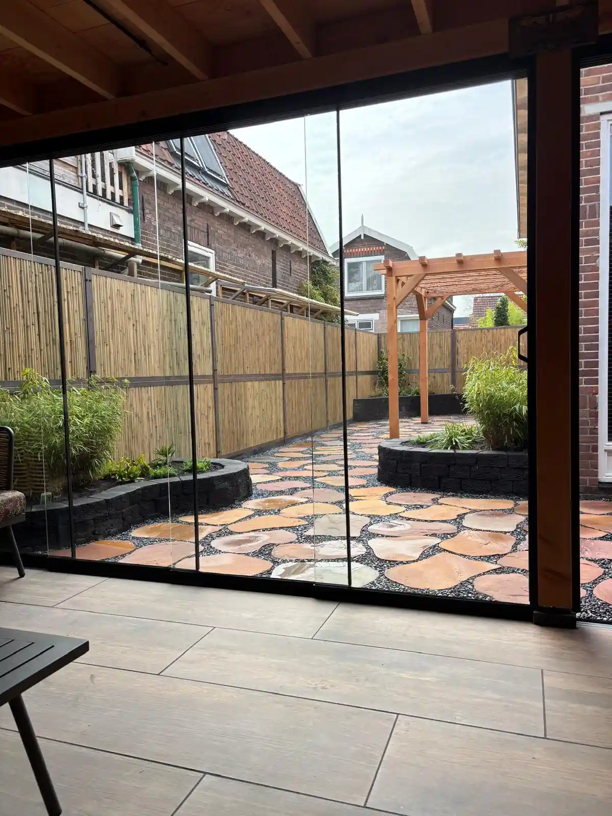 Zicht door de glazen schuifwand van een tuinkamer op een terras van flagstones in split, met bamboebeplanting in verhoogde borders en een Douglas pergola