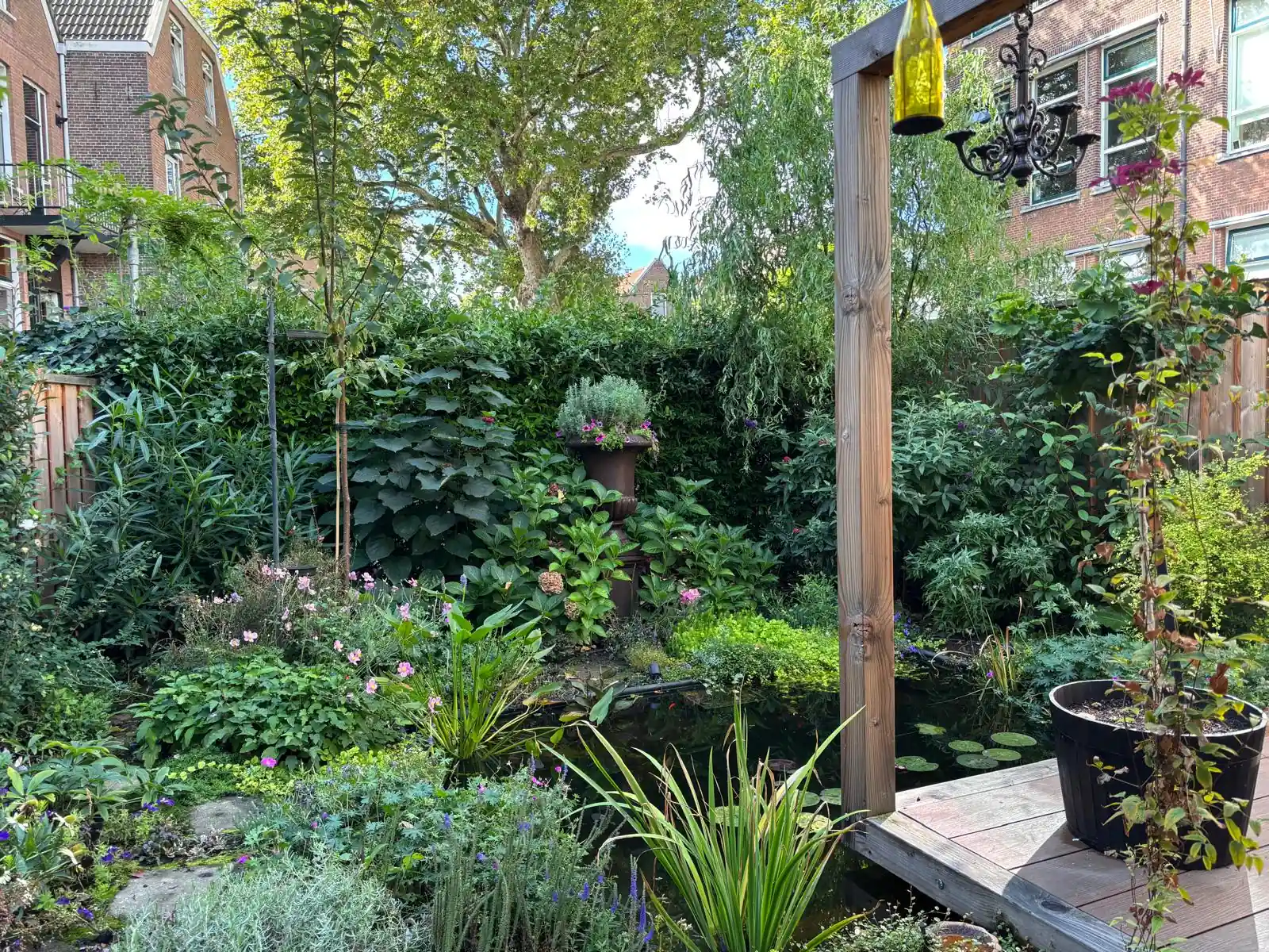 Vijver met waterlelies omringd door dichte borders met hortensia, herfstanemoon en siergrassen, met een klimopwand en een pergolapaal met glazen hanglamp