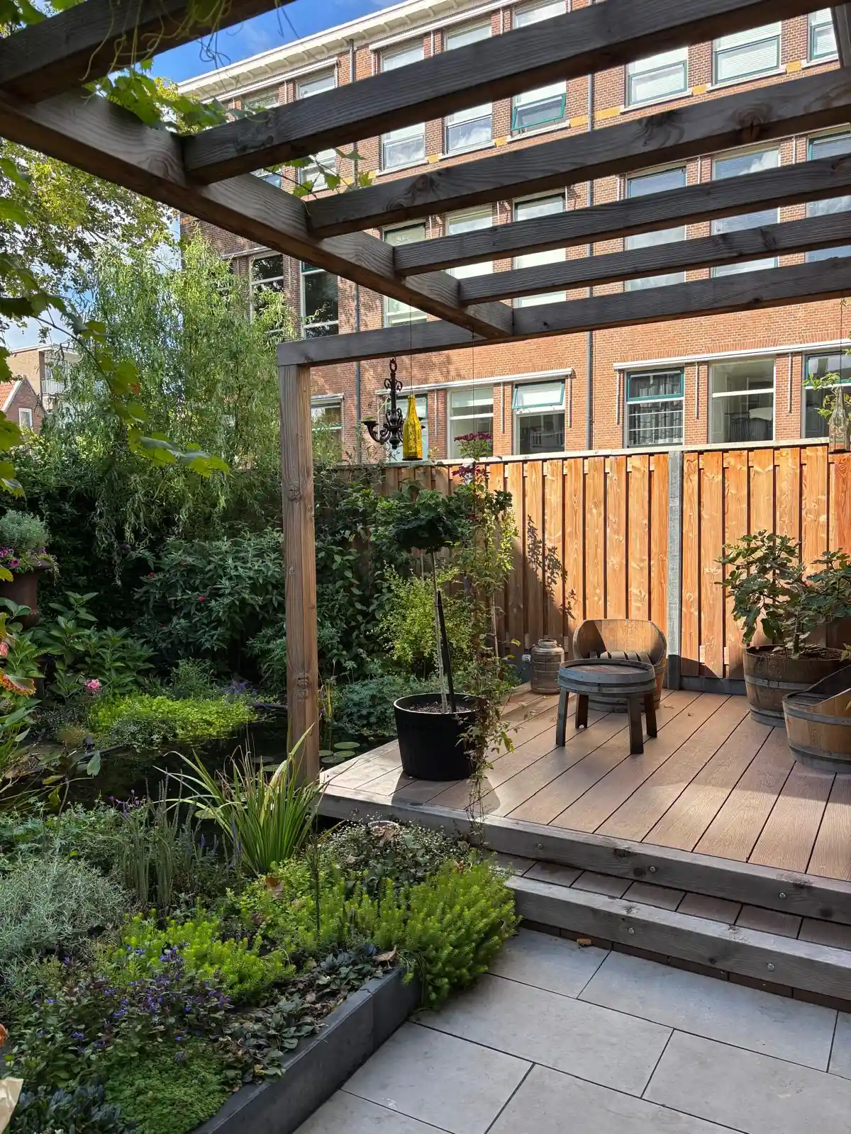 Verhoogd vlonderterras onder een pergola met stoelen en plantenbakken van oude wijnvaten, met een vijver en een border met sedum op de voorgrond