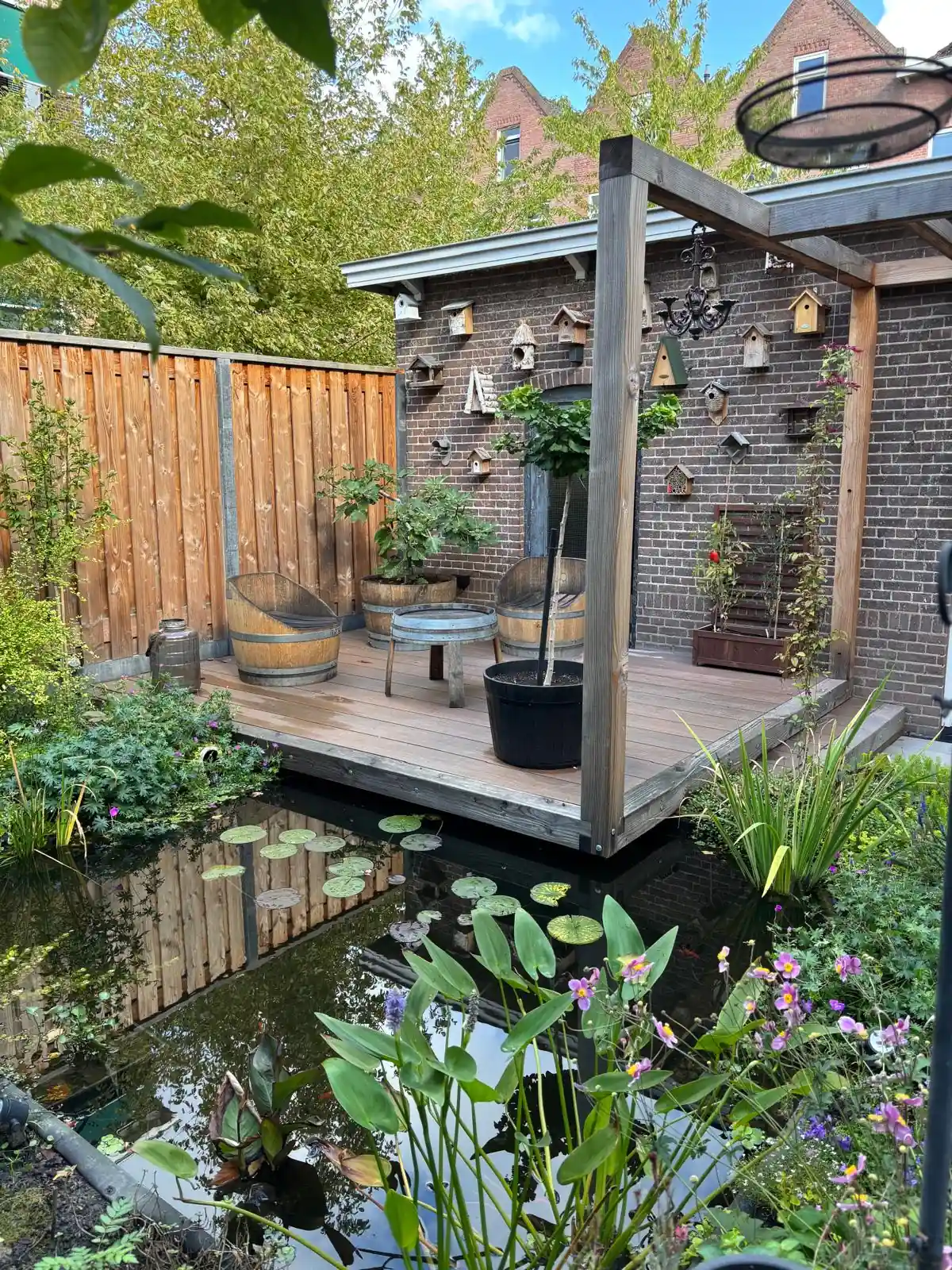 Vlonderterras aan de vijver met stoelen van wijnvaten, tegen een bakstenen gevel vol vogelhuisjes, met waterlelies en bloeiende oeverplanten in het water