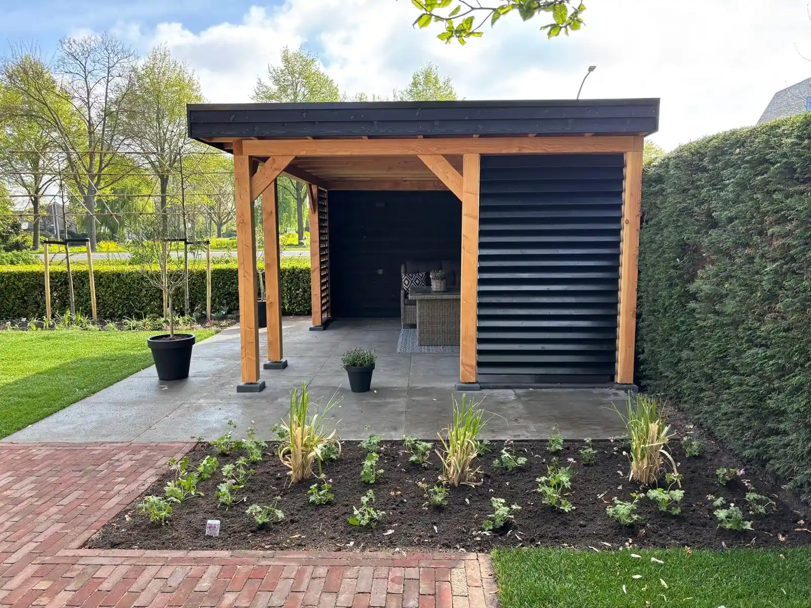 Overkapping met Douglas staanders en een zwarte lamellenwand op een terras van grote betontegels, met een verse border met ooievaarsbek en siergrassen ervoor