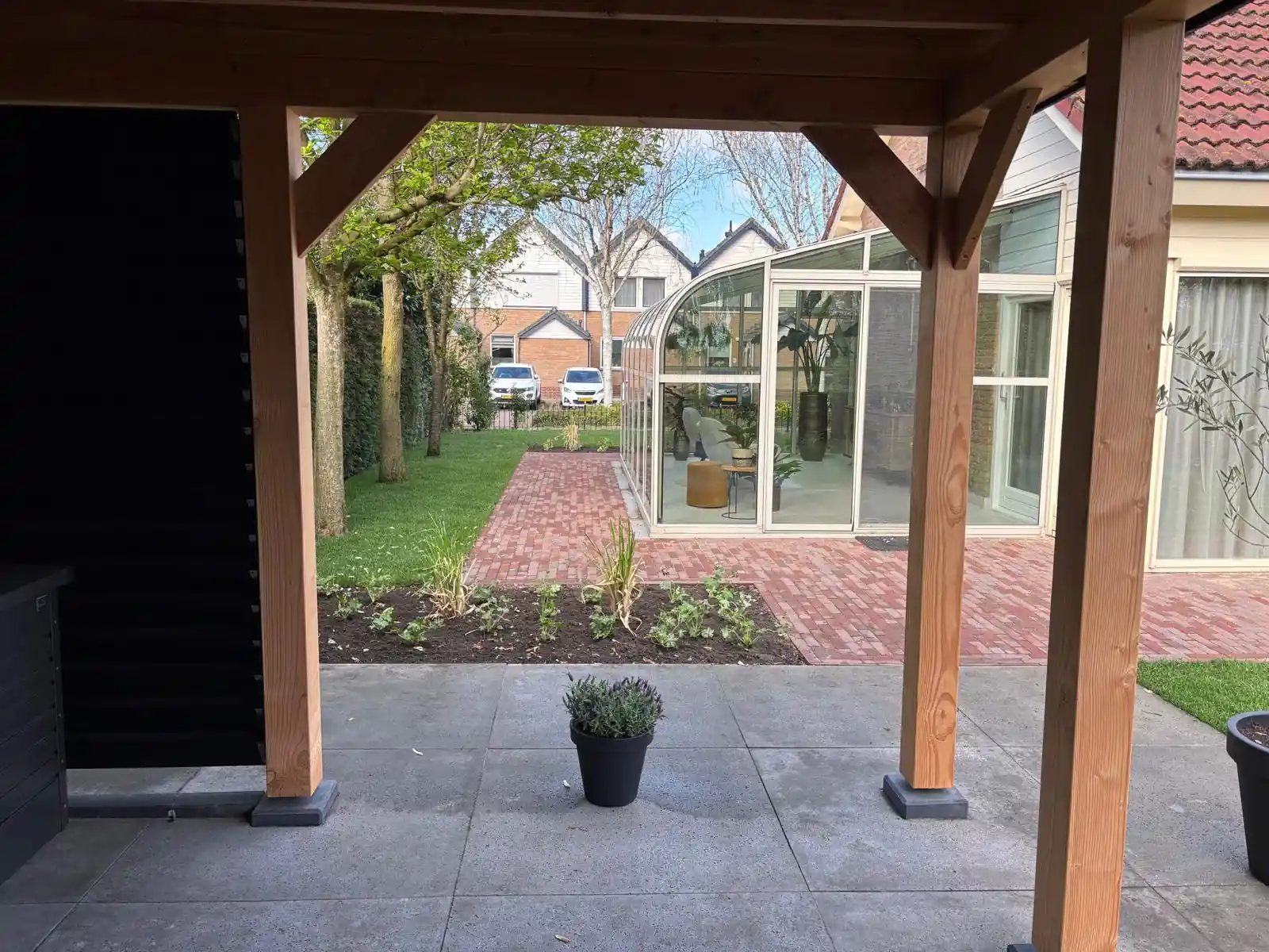 Zicht vanonder de overkapping op een gazon, een klinkerpad in visgraatverband en de gebogen glazen serre van de woning, met een pot lavendel op het terras