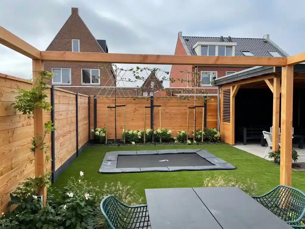 Rij jonge leibomen met een haag witte hortensia langs de schutting, met een verdiepte trampoline in het kunstgras en een pergola en overkapping ernaast