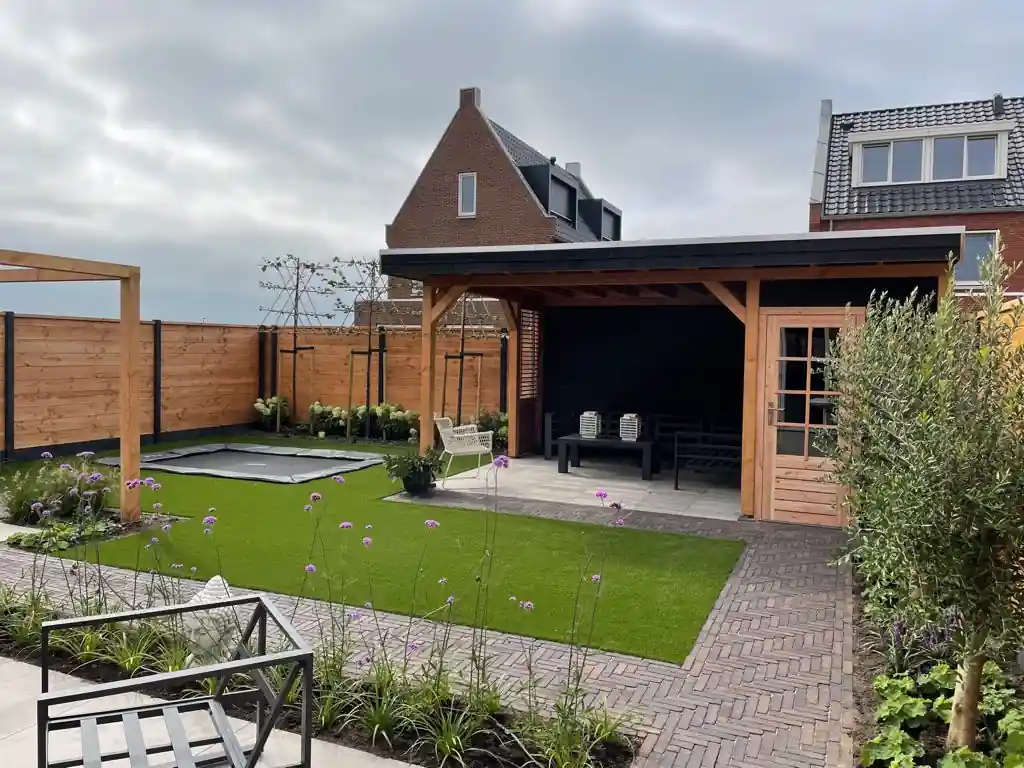 Houten overkapping met zwarte lamellenwand en loungebank, met daarvoor een veld kunstgras met verdiepte trampoline en een klinkerpad langs een olijfboom