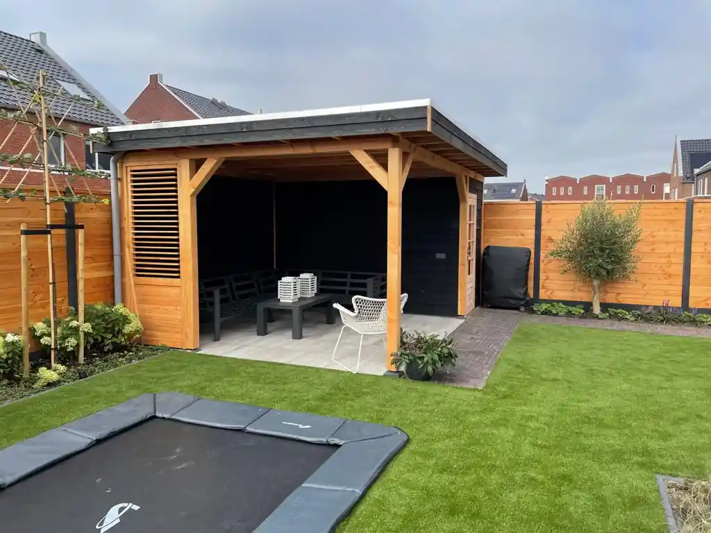Houten overkapping met lamellenwand en donkere loungebank op een vloer van lichte planktegels, met een klinkerpad, olijfboom en verdiepte trampoline in het kunstgras