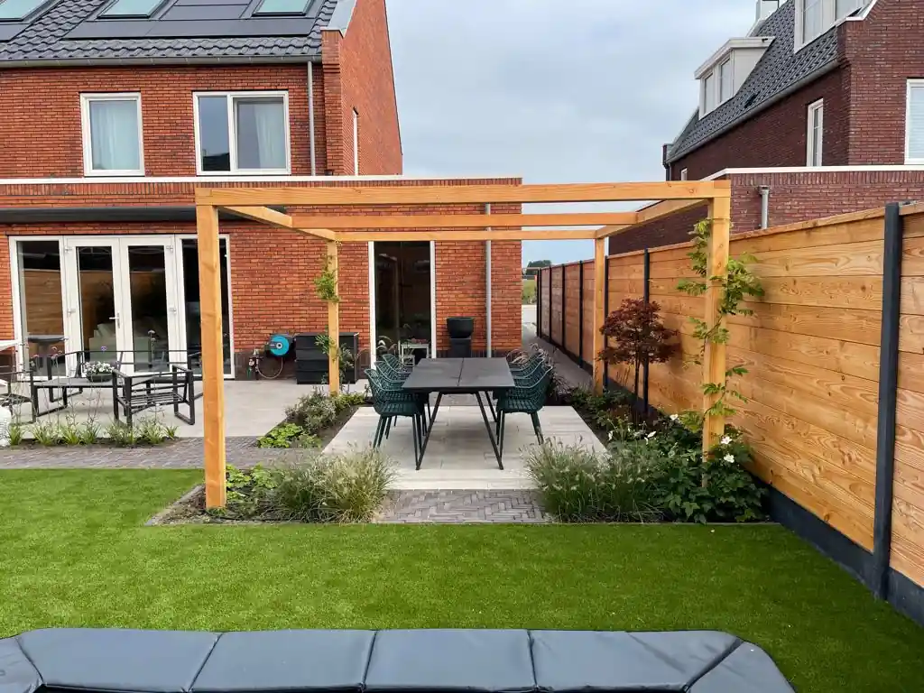 Douglas pergola boven een terras van lichte tegels voor de openslaande deuren van een bakstenen woning, met siergrassen, klimplanten en kunstgras