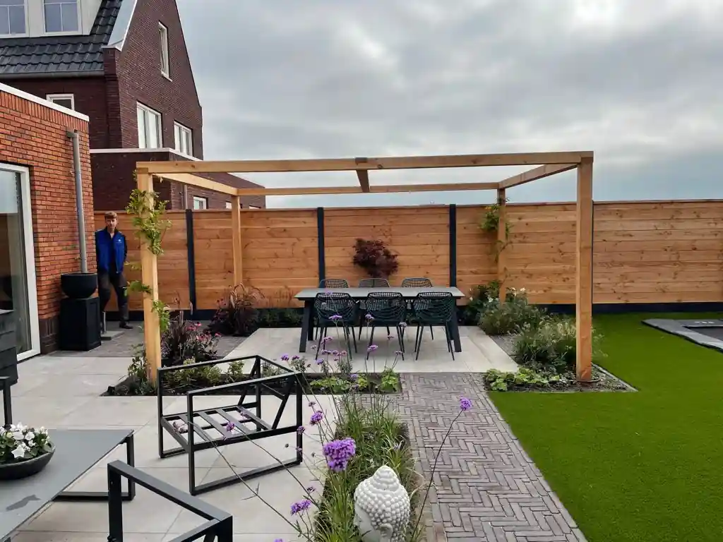 Douglas pergola boven een eettafel met zwarte draadstoelen op een terras van lichte tegels, met borders vol paars bloeiend ijzerhard en een klinkerpad in visgraatverband