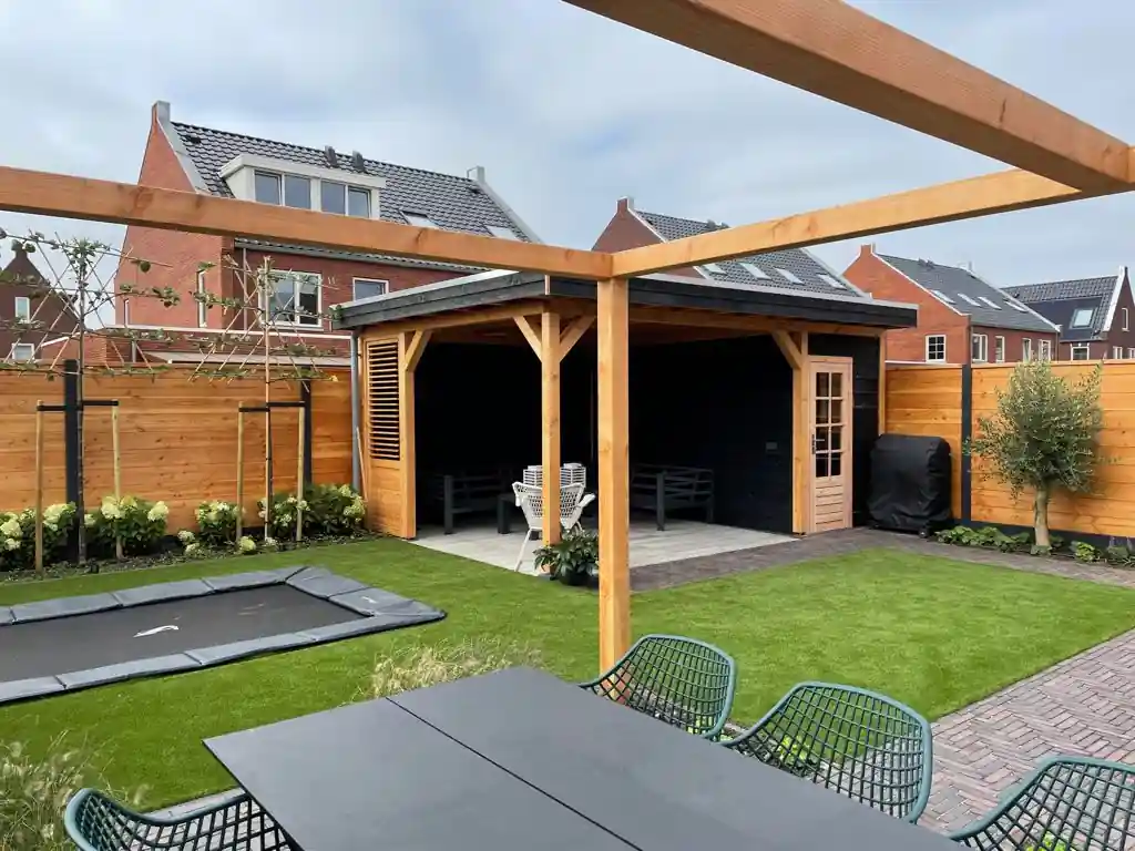 Overzicht met op de voorgrond een Douglas pergola boven de eethoek en daarachter een overkapping met loungebank, kunstgras, trampoline en een olijfboom