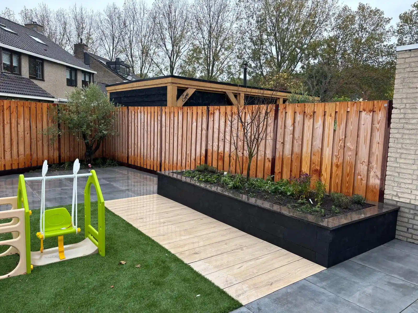 Langgerekte verhoogde border van zwarte stapelblokken met jonge beplanting langs een pad van houtlook tegels, met kunstgras, speeltoestel en een olijfboom