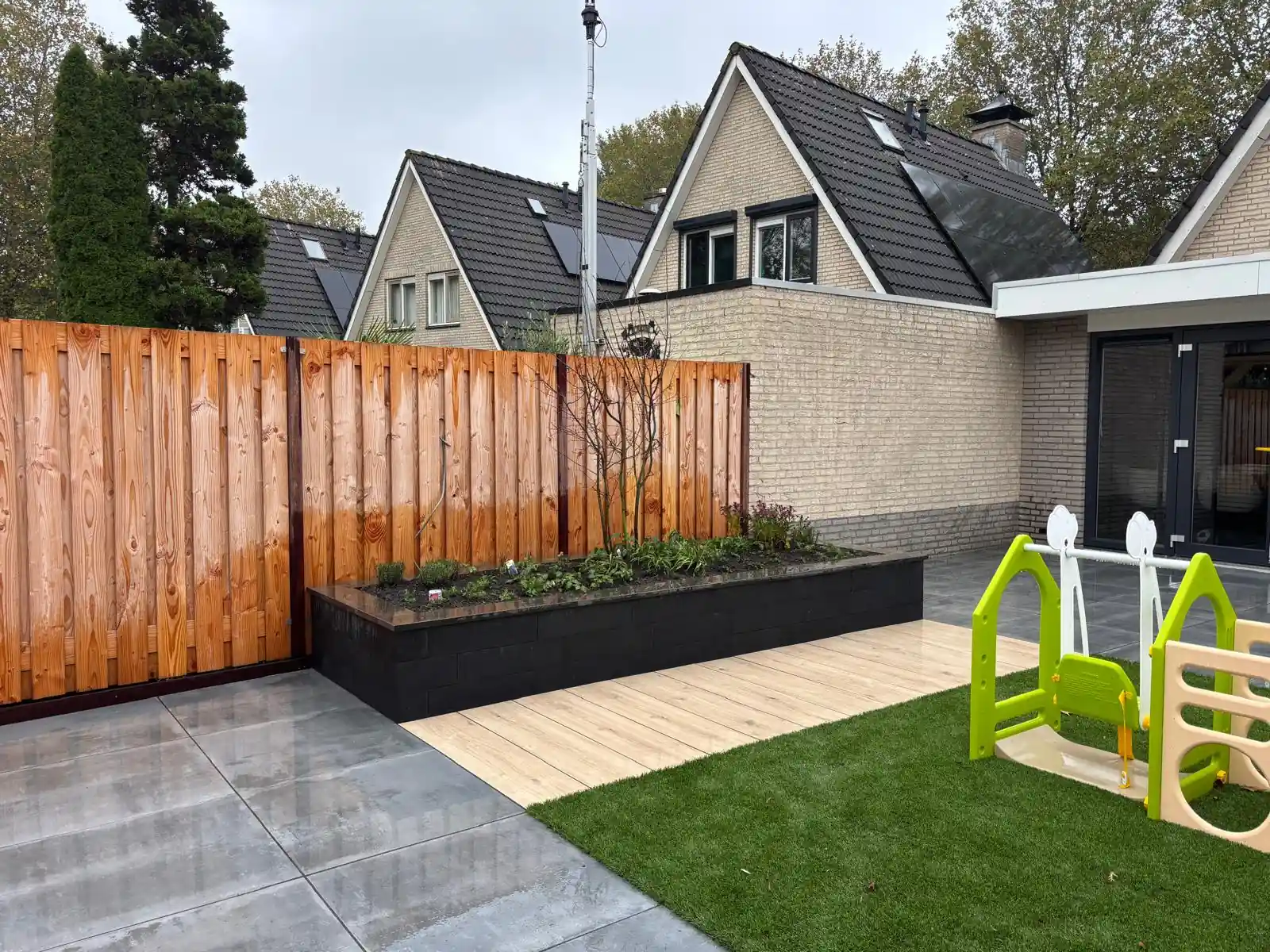 Veld kunstgras met speeltoestel grenzend aan een pad van houtlook tegels en een verhoogde border, met de lichte bakstenen gevel van de woning erachter